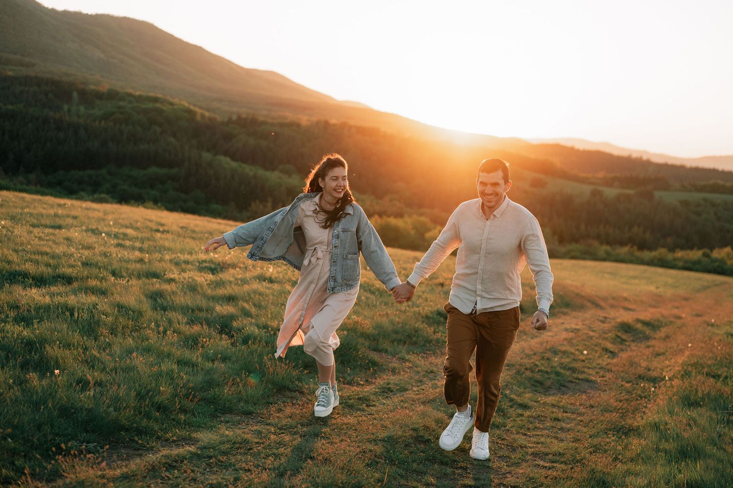 Two people run playfully through a grassy field at sunset, creating a romantic silhouette against the golden sky.