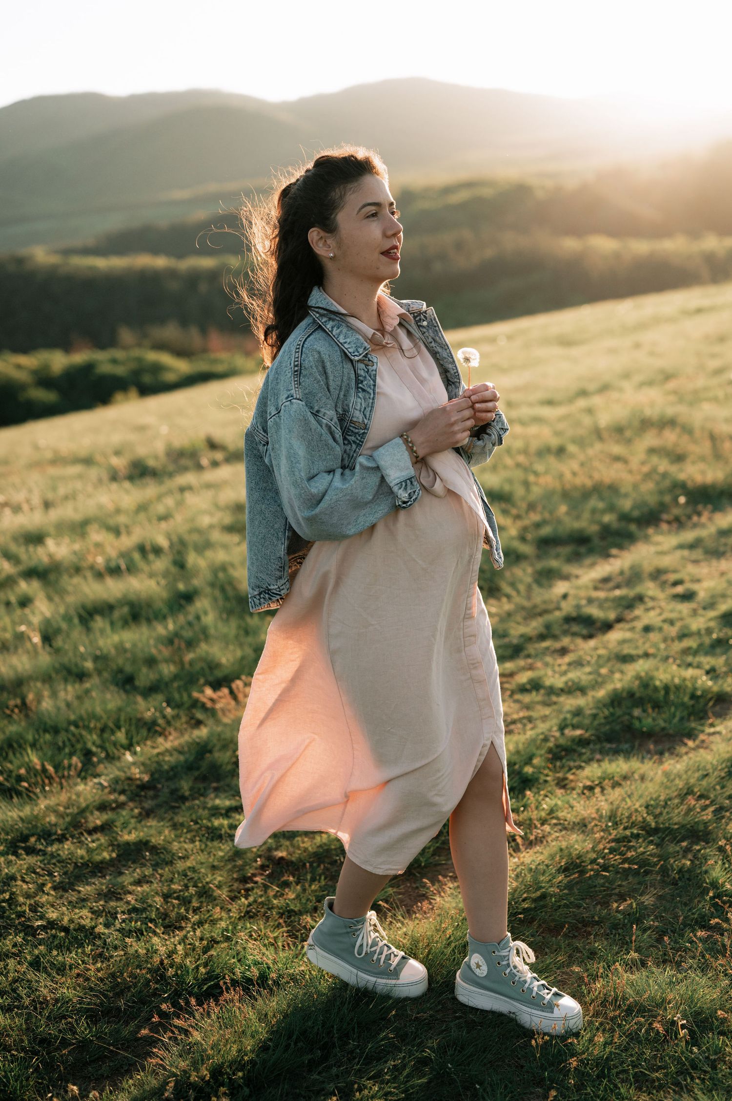 Person in denim jacket and white dress sits on grassy hillside during golden hour sunset.