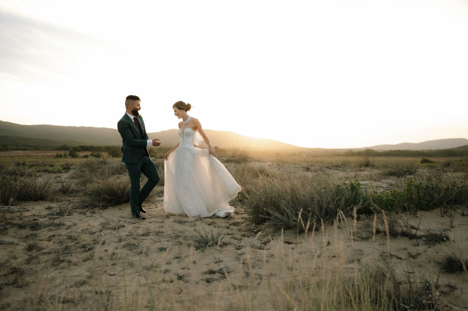 Bride and groom walk hand in hand across a desert landscape at golden sunset with mountains in the background.