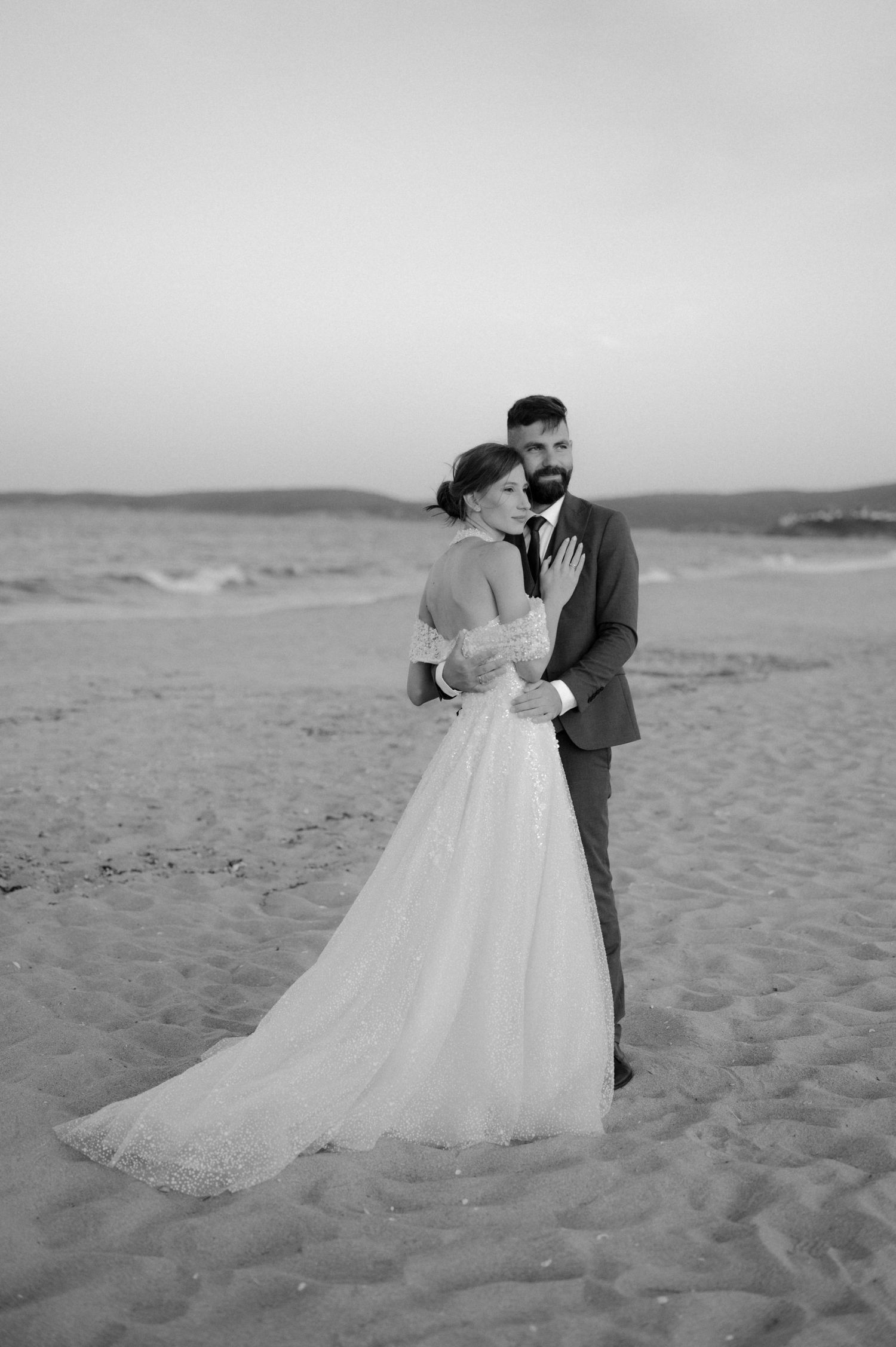 Black and white photo of a bride and groom embracing on a sandy beach, with ocean waves in the background.