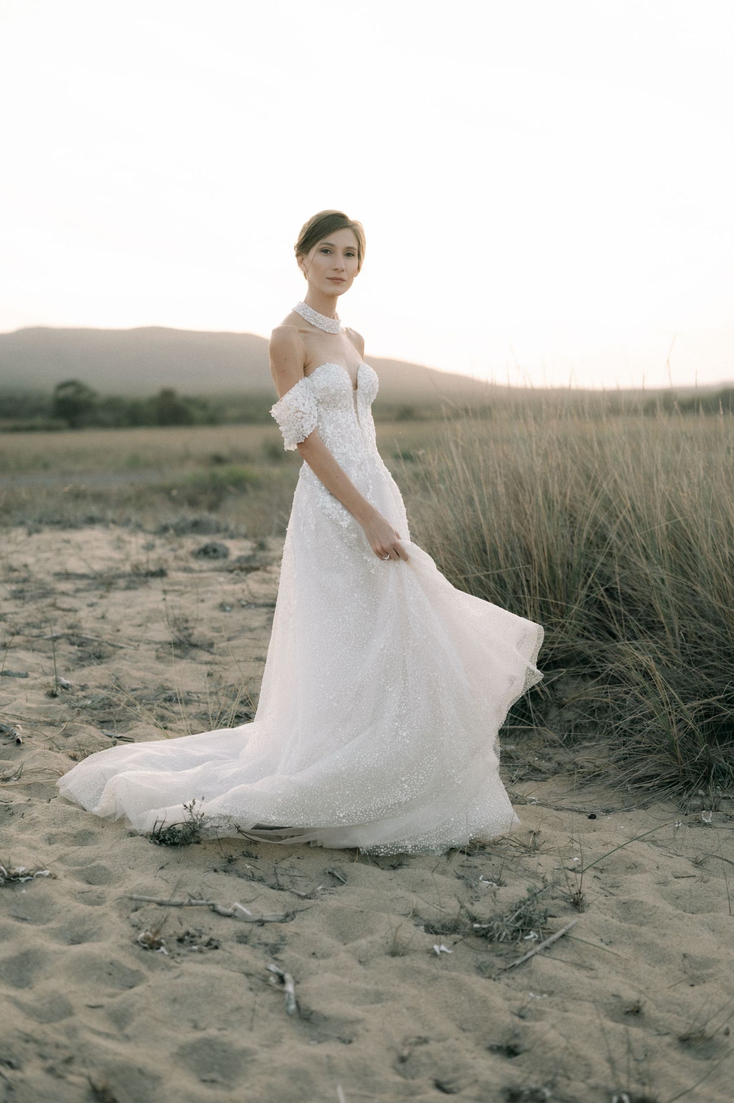 Bride in off-shoulder white lace wedding gown with flowing train poses on sandy beach at sunset.