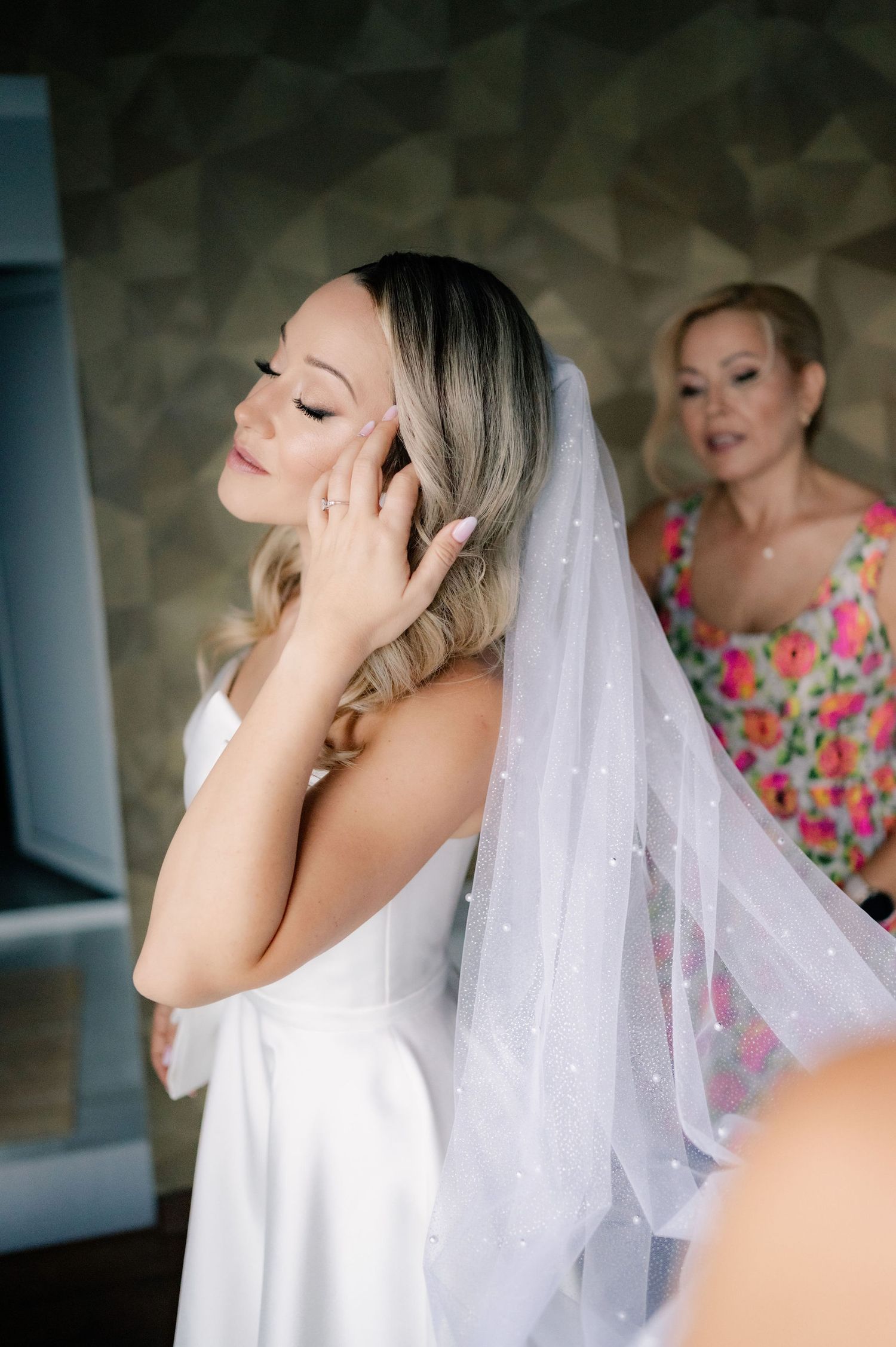 A bride in a white dress and veil getting ready on her wedding day with someone helping in a floral dress behind her.
