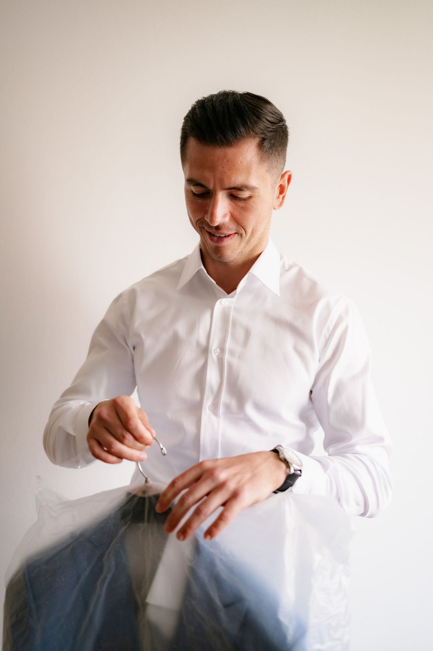 A person wearing a crisp white dress shirt and watch adjusts their sleeve while looking down with a smile.