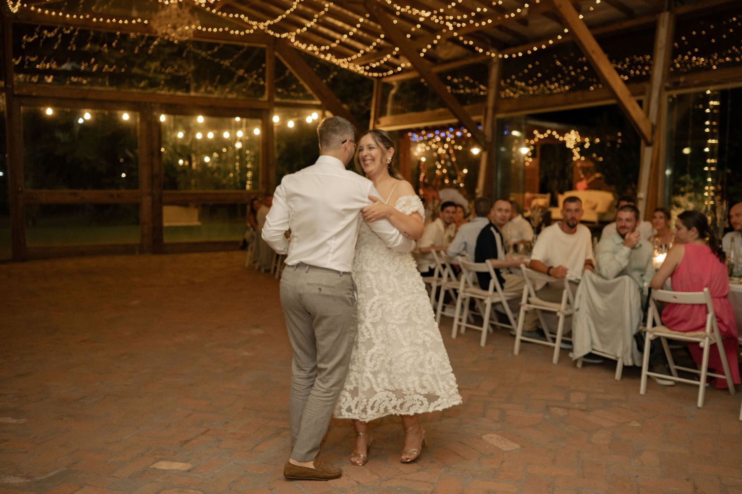 Wedding guests dance together on a brick patio under twinkling string lights at an outdoor evening reception.