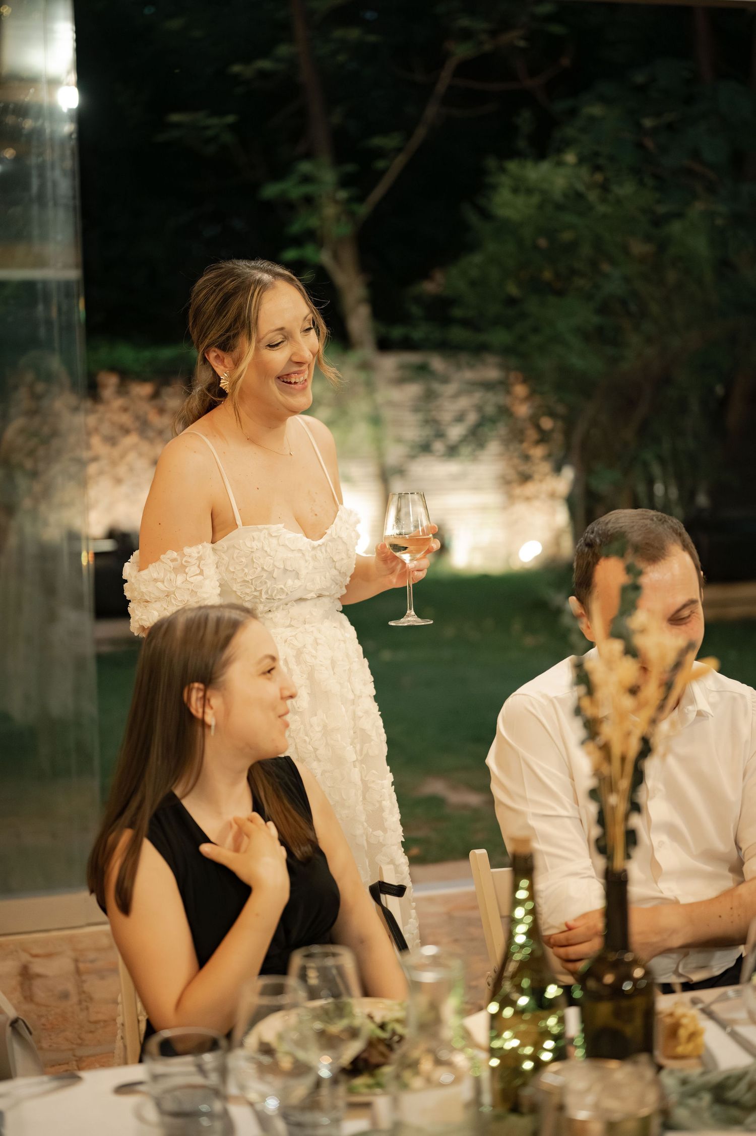 Wedding guests share laughter and wine during an intimate outdoor evening reception with twinkling lights.