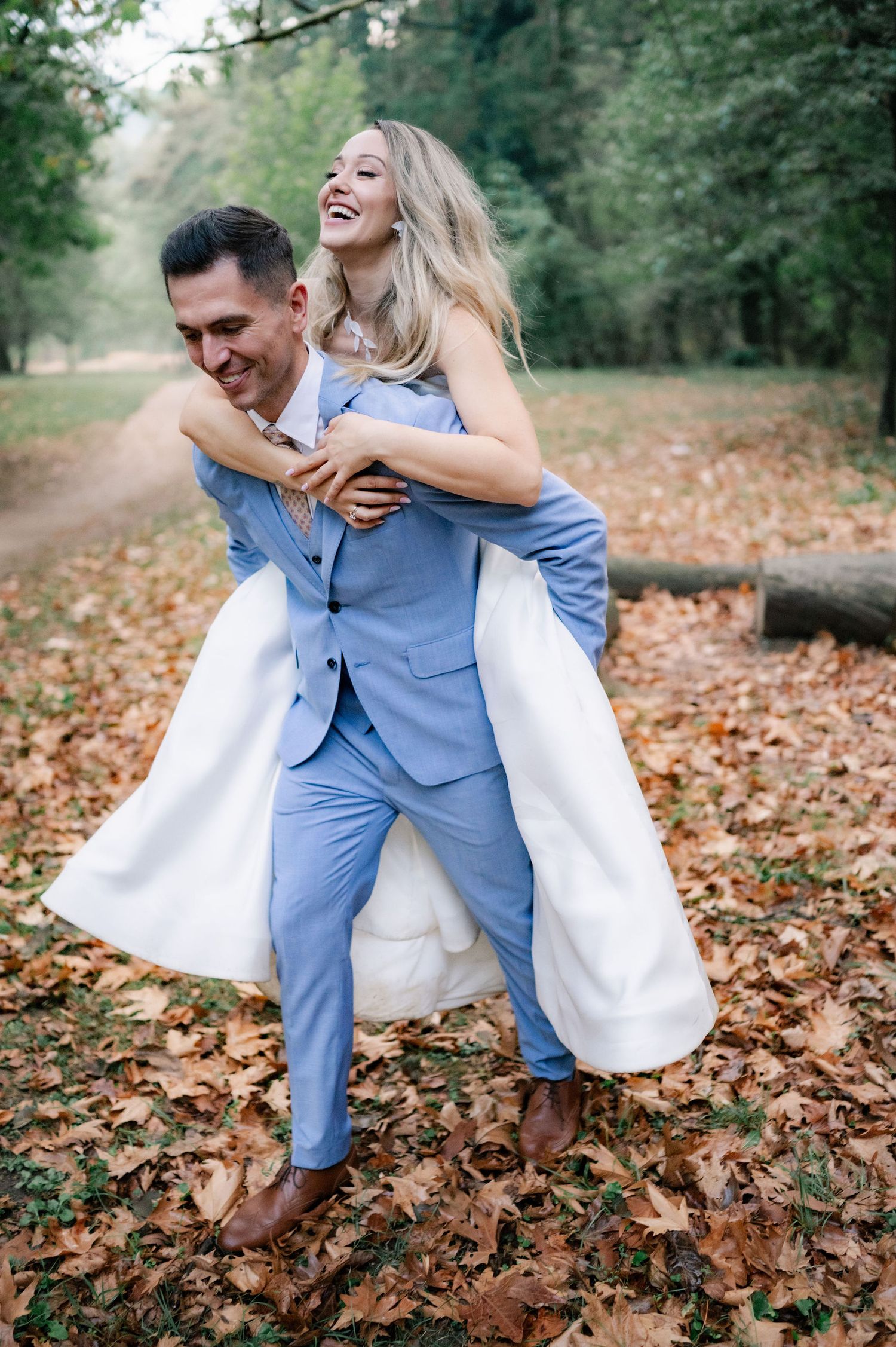 A joyful couple shares a playful piggyback ride moment on an autumn path covered in fallen leaves.