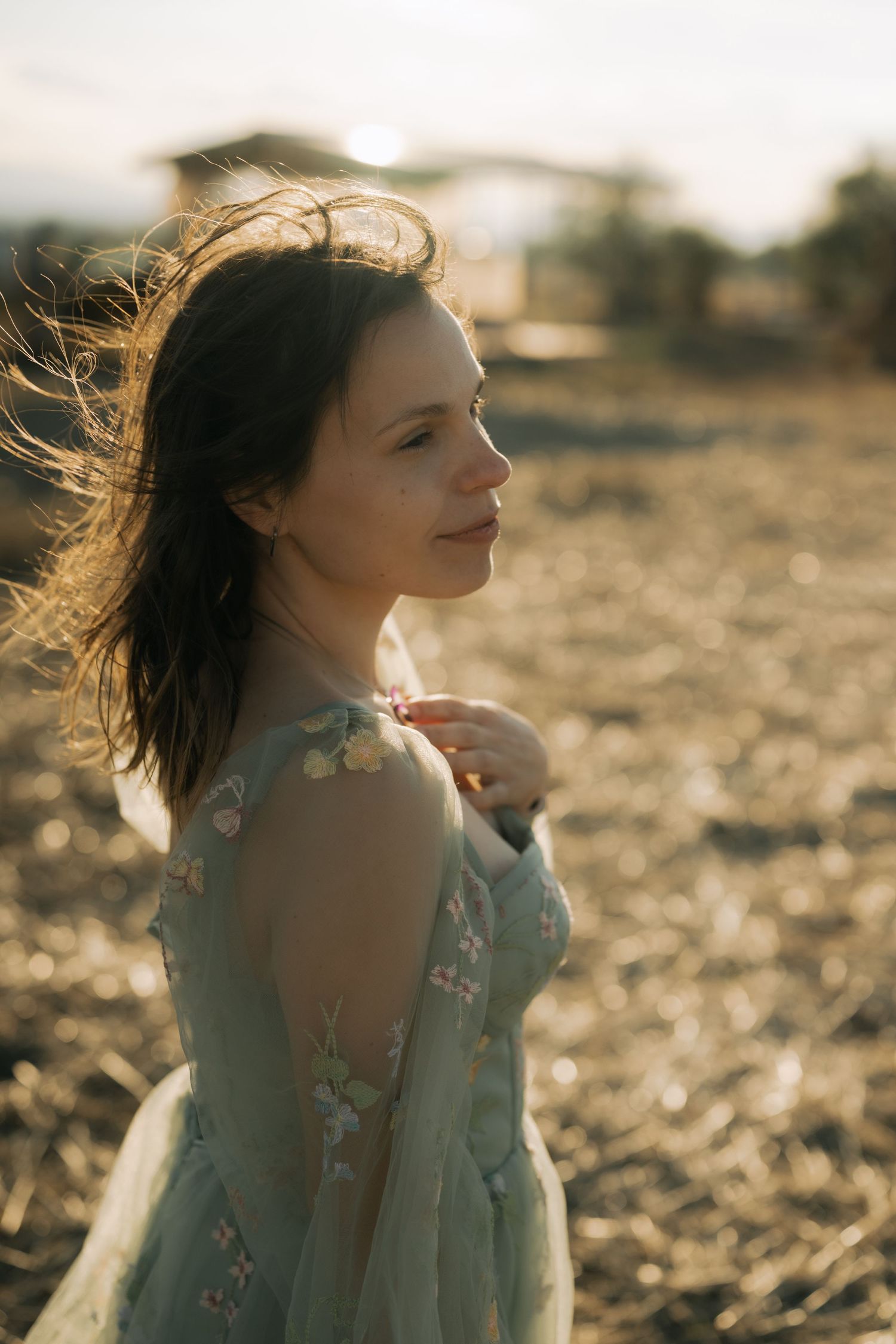 Person in flowy dress poses in golden sunlight with windswept hair against natural outdoor backdrop.