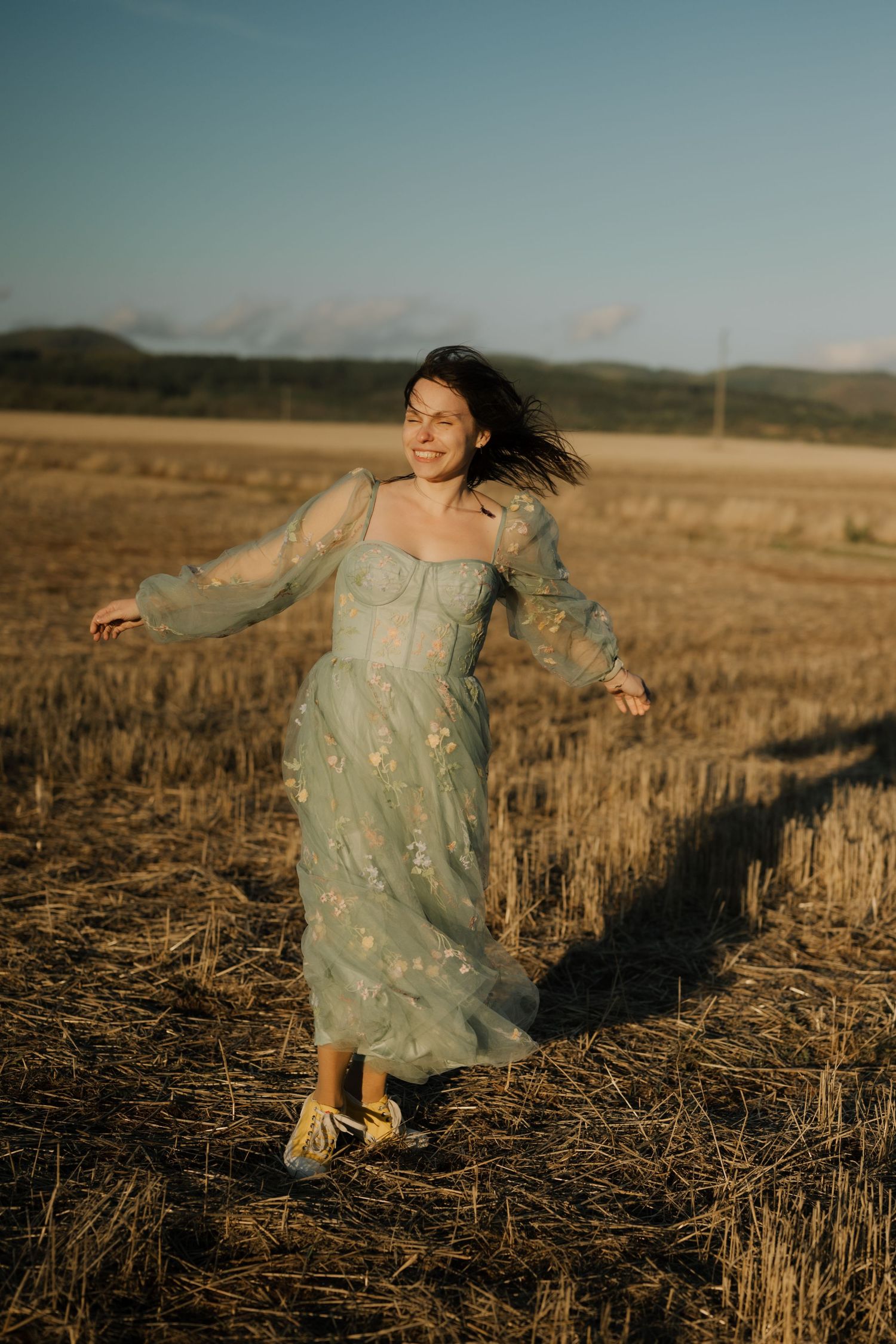 A figure in a flowy mint dress dances joyfully through a golden harvested field at sunset.