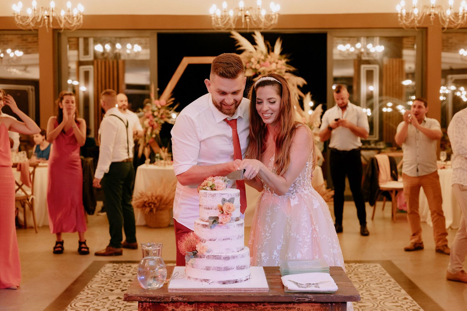 Wedding couple cuts their elegant white tiered cake at an ornate reception hall with crystal chandeliers.