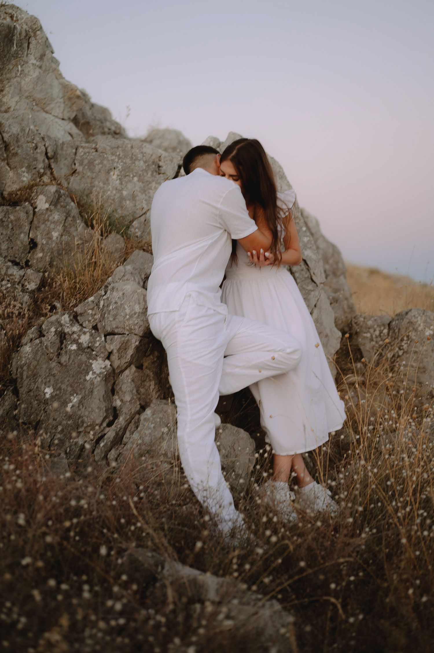 A romantic couple in white outfits embrace against a rocky cliff backdrop during sunset.