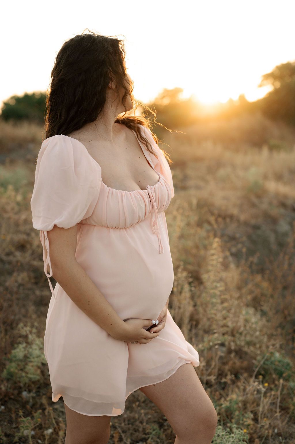 A pregnant woman in a flowing white dress stands in a golden field at sunset, her hair gently blowing in the breeze.