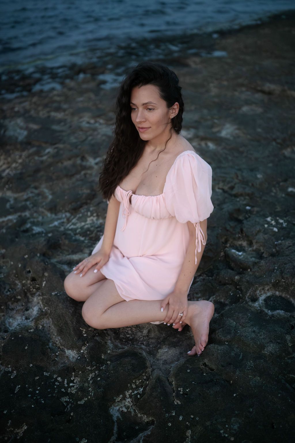 A person in a light pink dress poses gracefully on dark rocky terrain during a moody outdoor photoshoot.