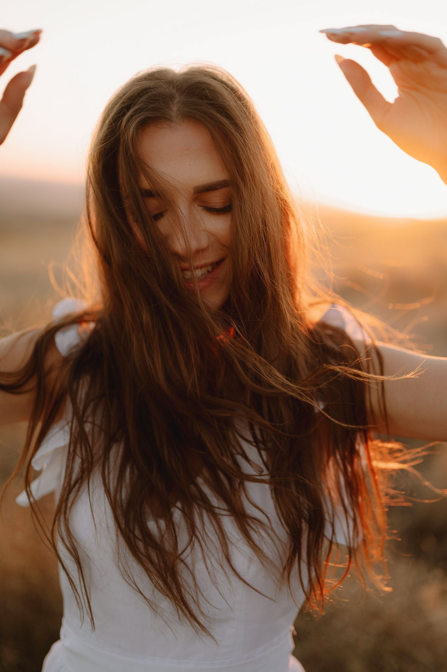 Long brown hair flows in the golden sunlight as a woman in white smiles with closed eyes during sunset.