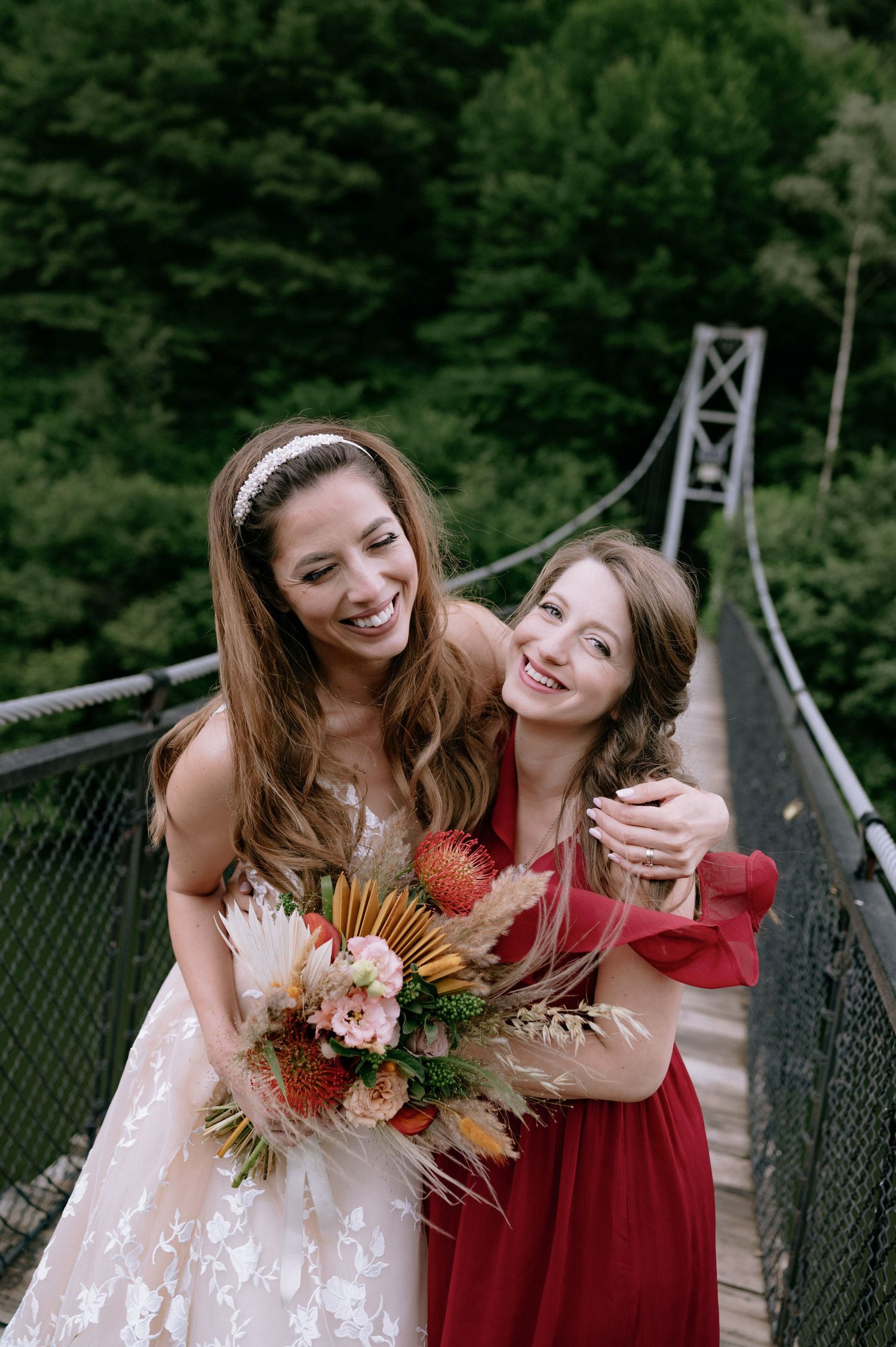 Two people in white and burgundy dresses share joyful moments together on a suspension bridge surrounded by trees.