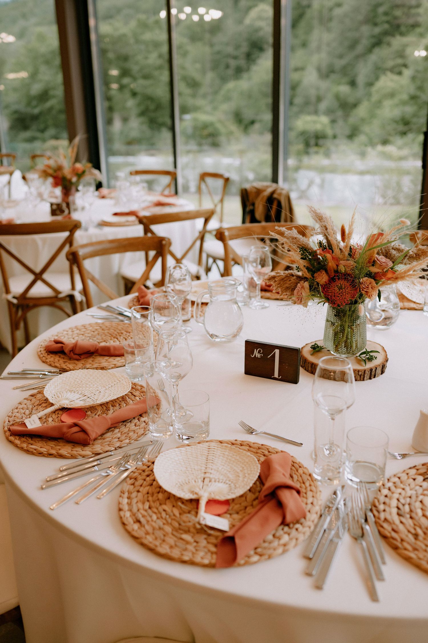 Elegant rustic wedding table setting with woven placemats, coral napkins, and fall floral centerpiece on white tablecloth.