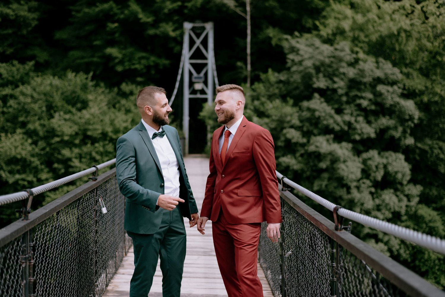 Two people in formal attire share a romantic moment on a wooden suspension bridge in a lush forest setting.