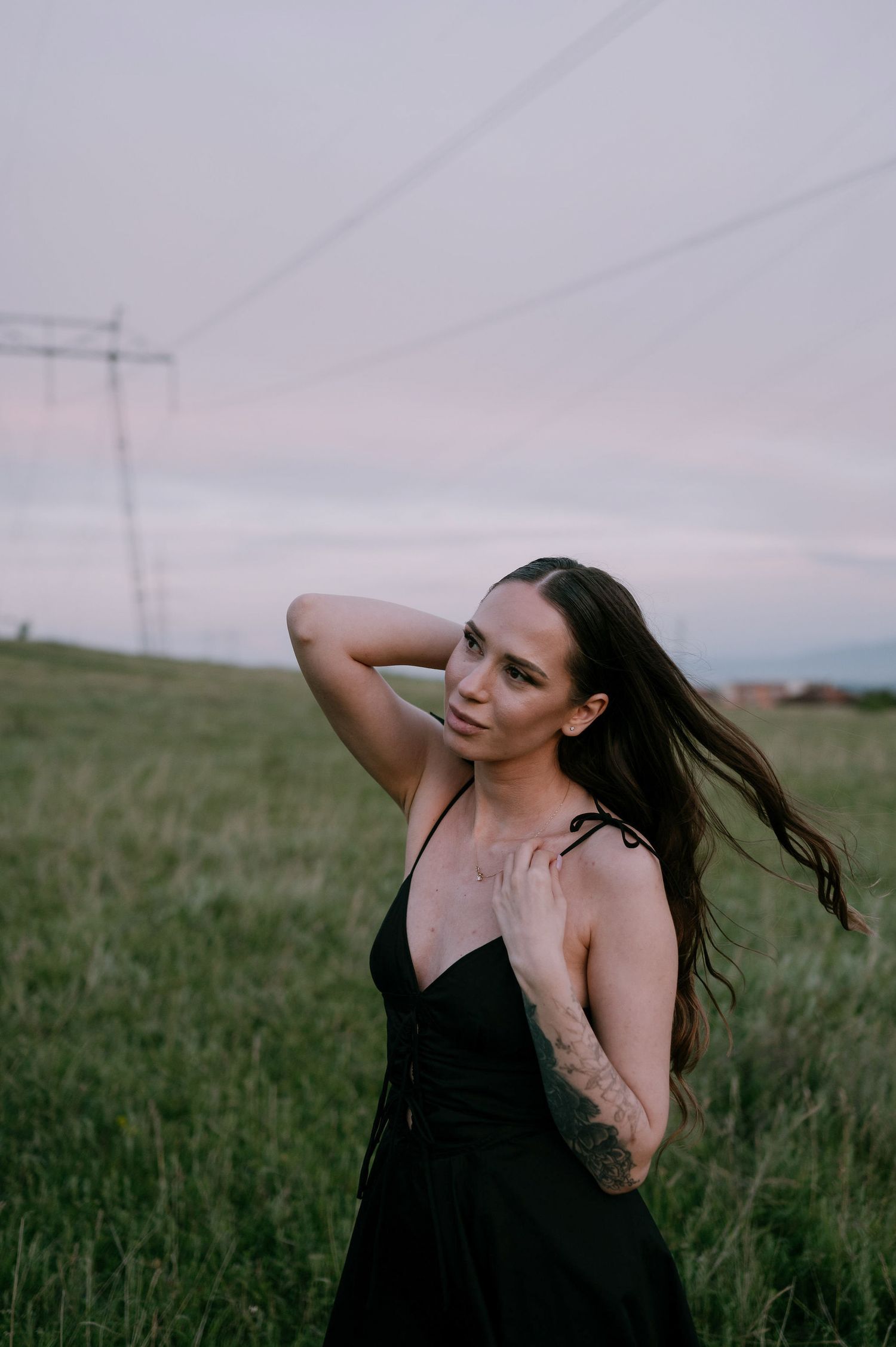 Woman in black dress poses gracefully in a grassy field during sunset, with power lines visible in the background.