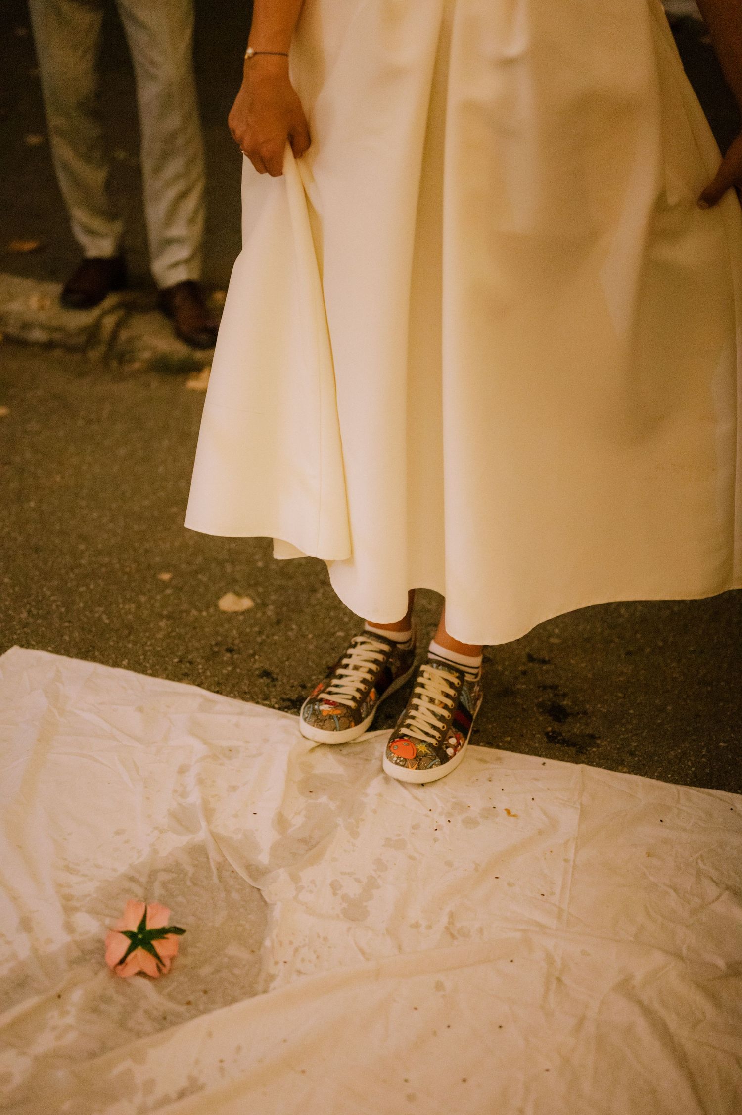 A pair of sneakers peek out from under a long white dress on a white fabric-covered surface.
