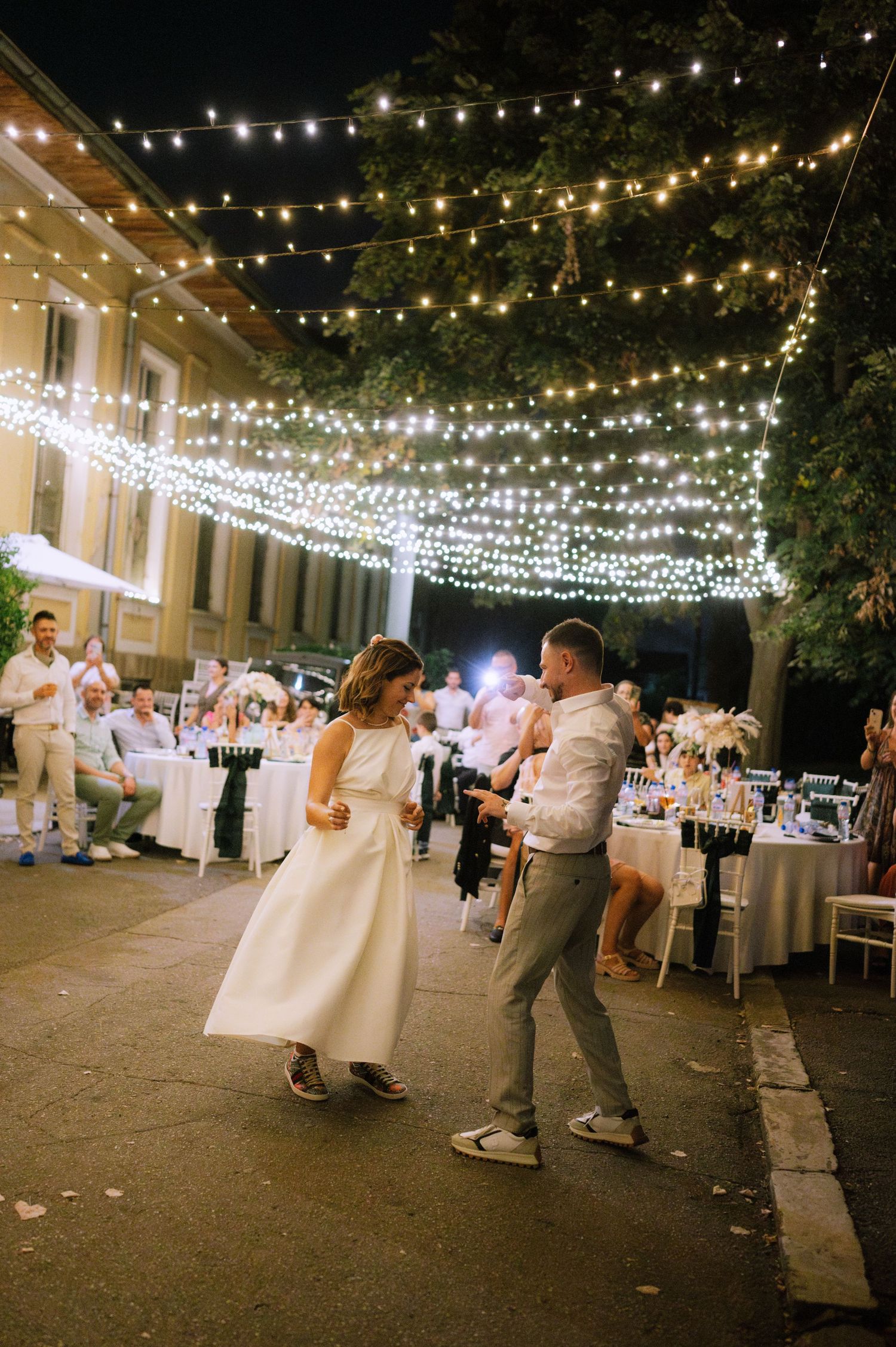 Wedding guests dance under twinkling string lights at an outdoor evening reception.