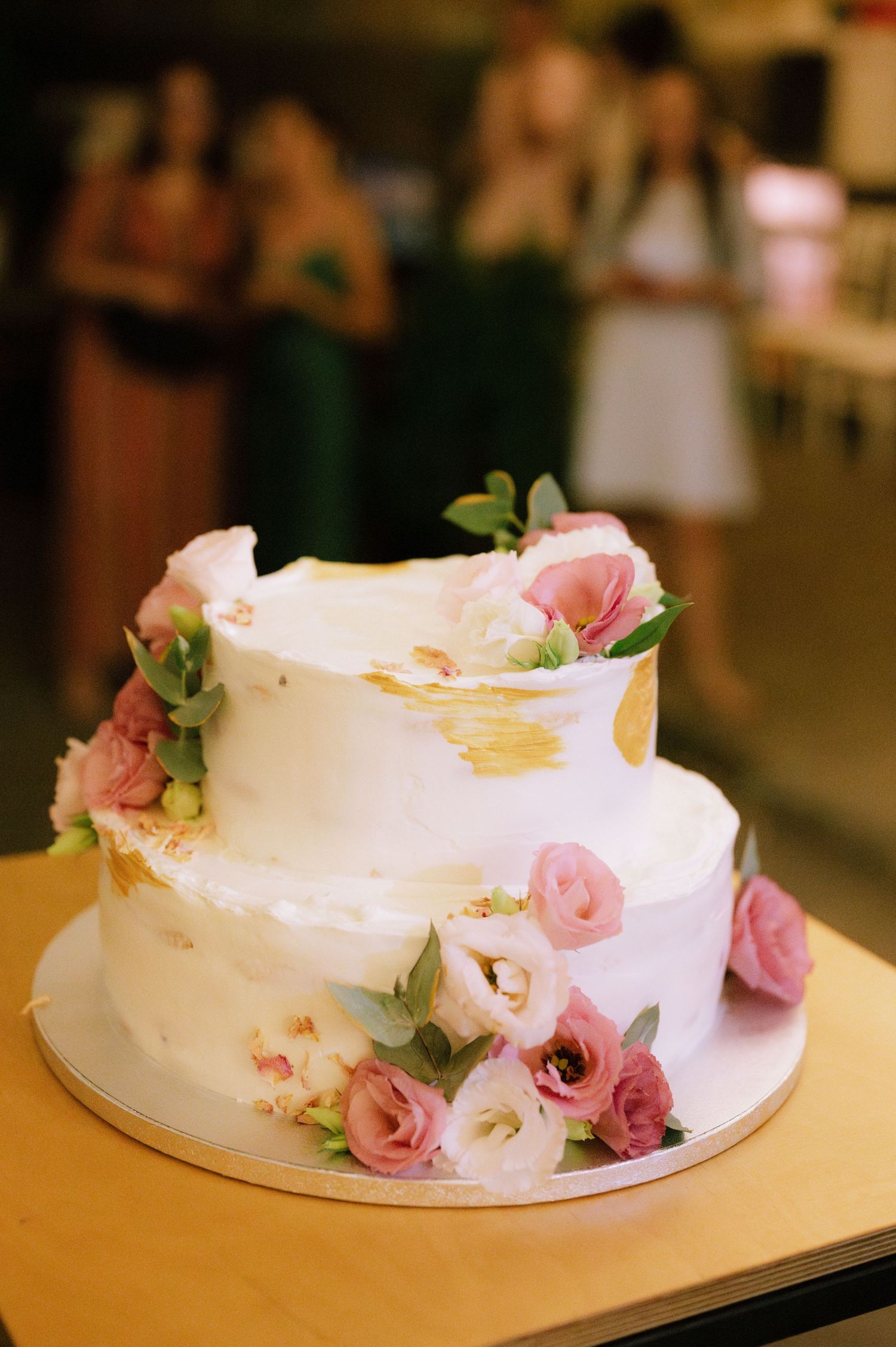 A two-tiered white wedding cake decorated with delicate pink roses and gold accents on a yellow surface.