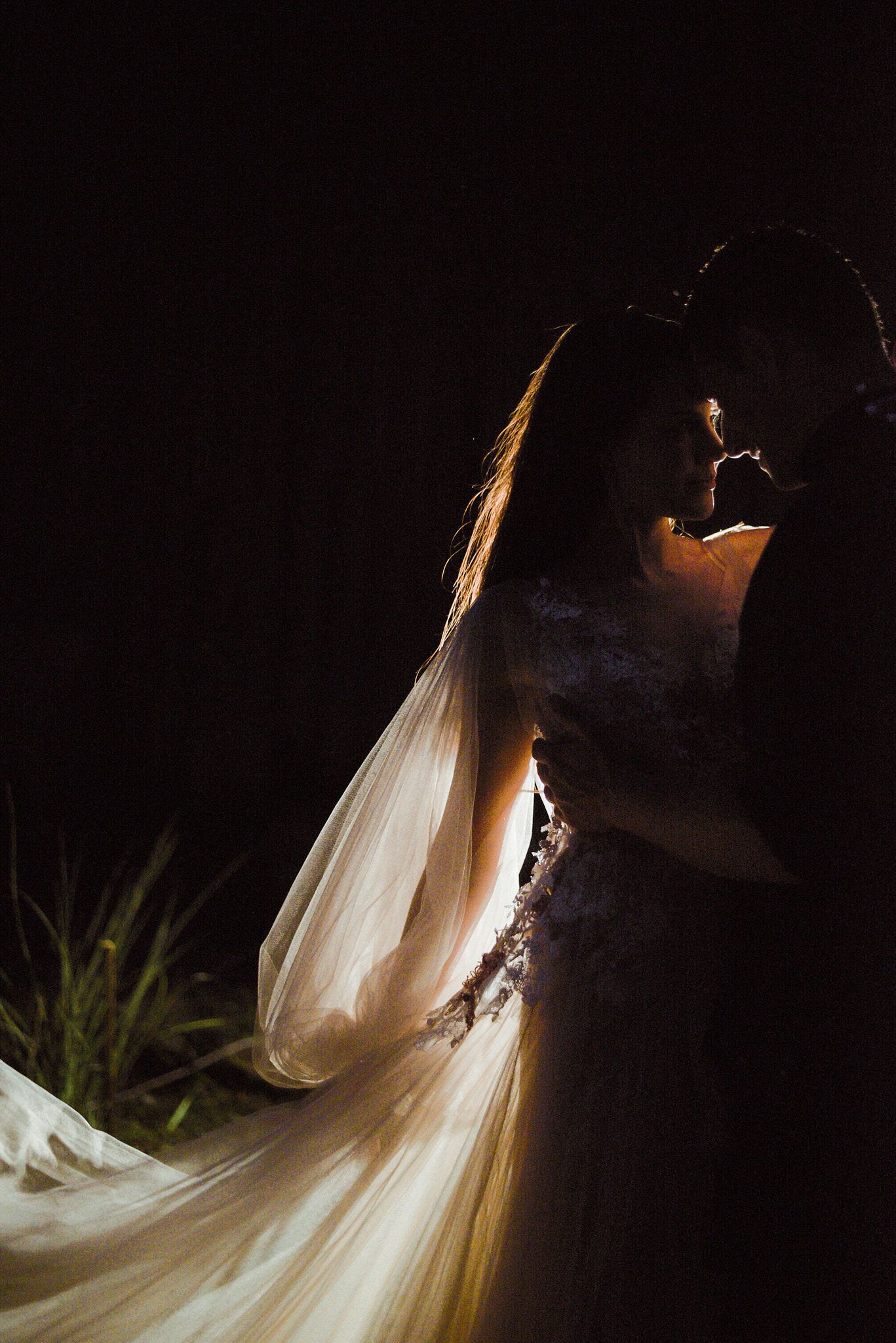A flowing white wedding dress veil creates an ethereal effect against a dark background in a dramatic bridal photo series.