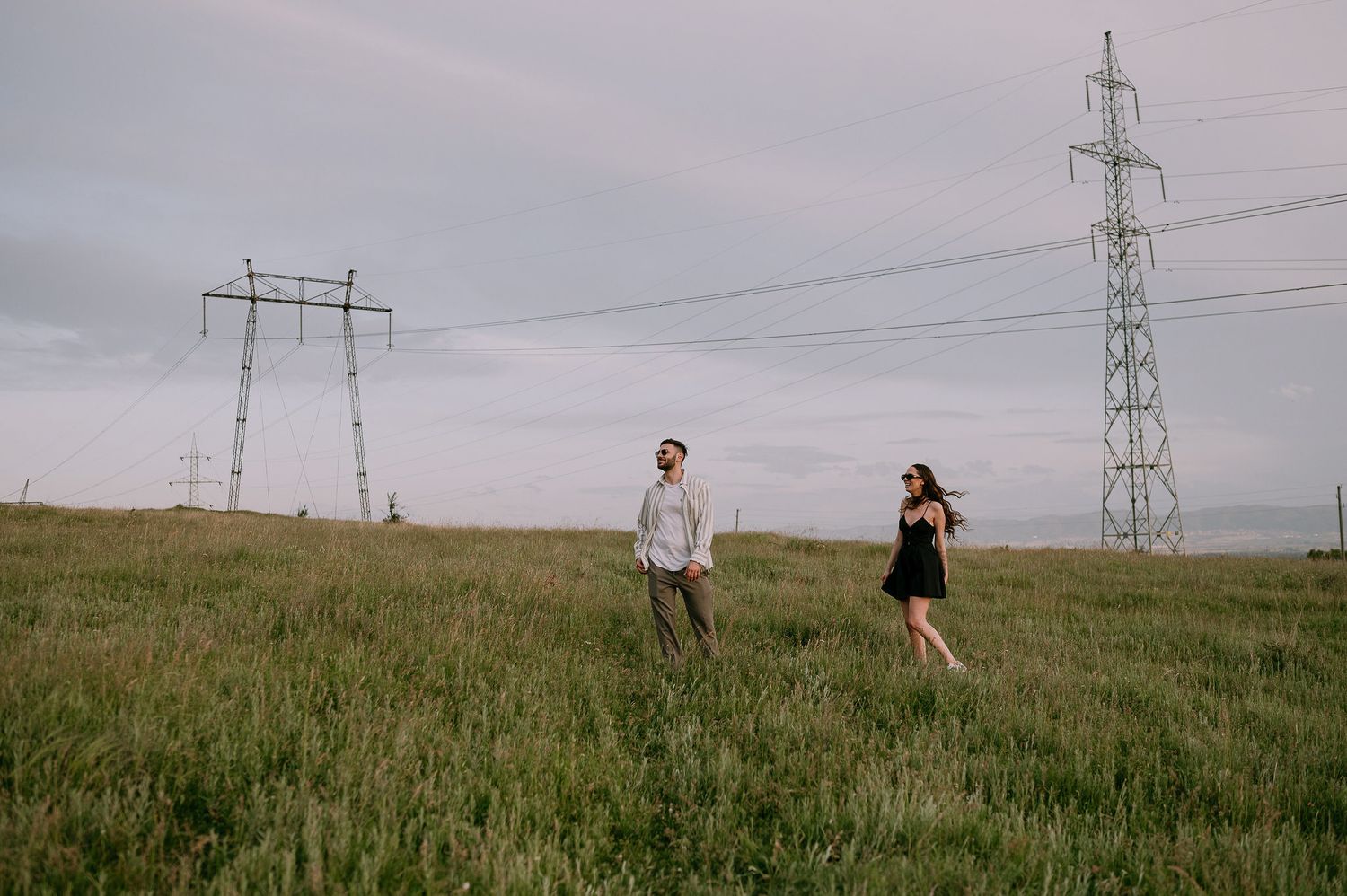 People walking on a grassy hill with electrical transmission towers and power lines in the background under cloudy skies.