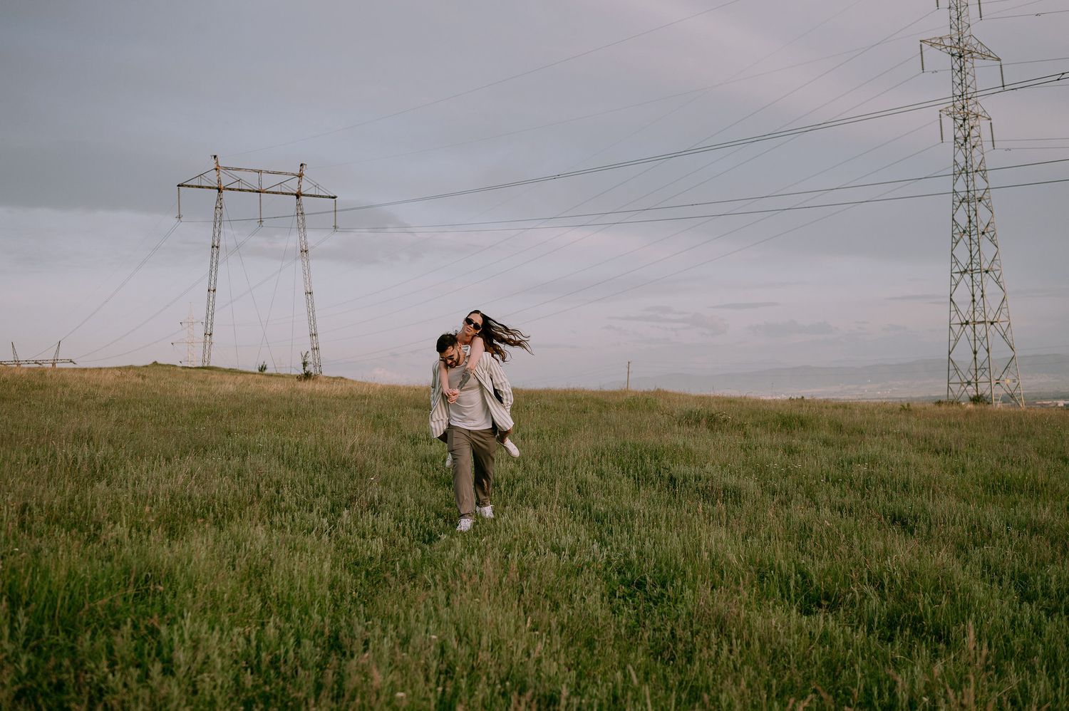 Person standing in a grassy field beneath electrical power lines and towers on an overcast day.