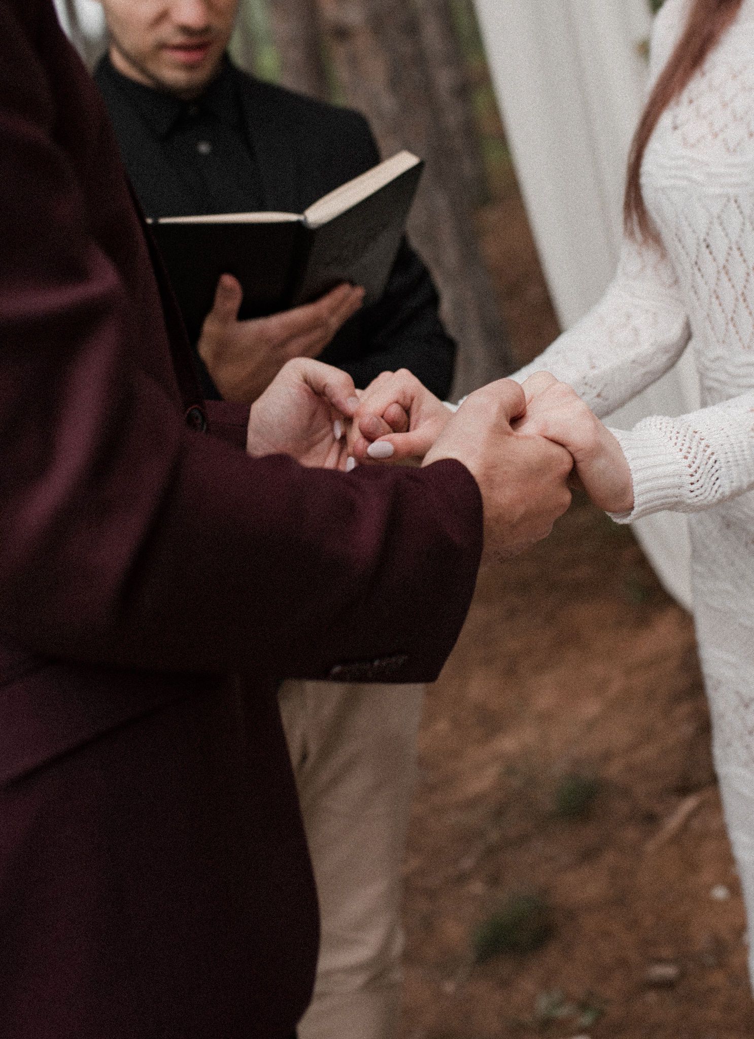 A couple exchanges rings during an intimate outdoor wedding ceremony, focusing on their hands and formal attire.