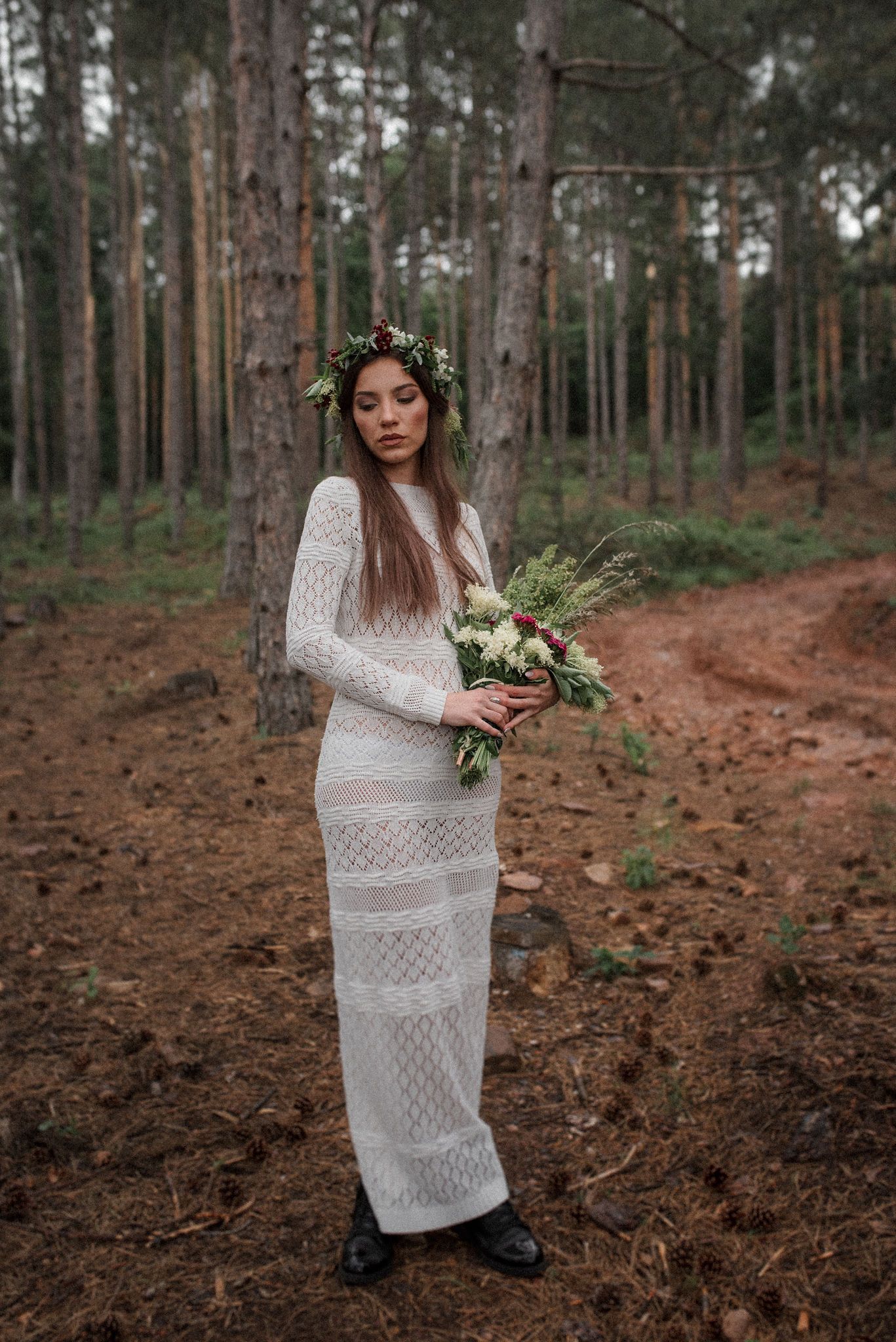 A person in a white lace dress holds a floral bouquet while standing among tall pine trees in a forest.