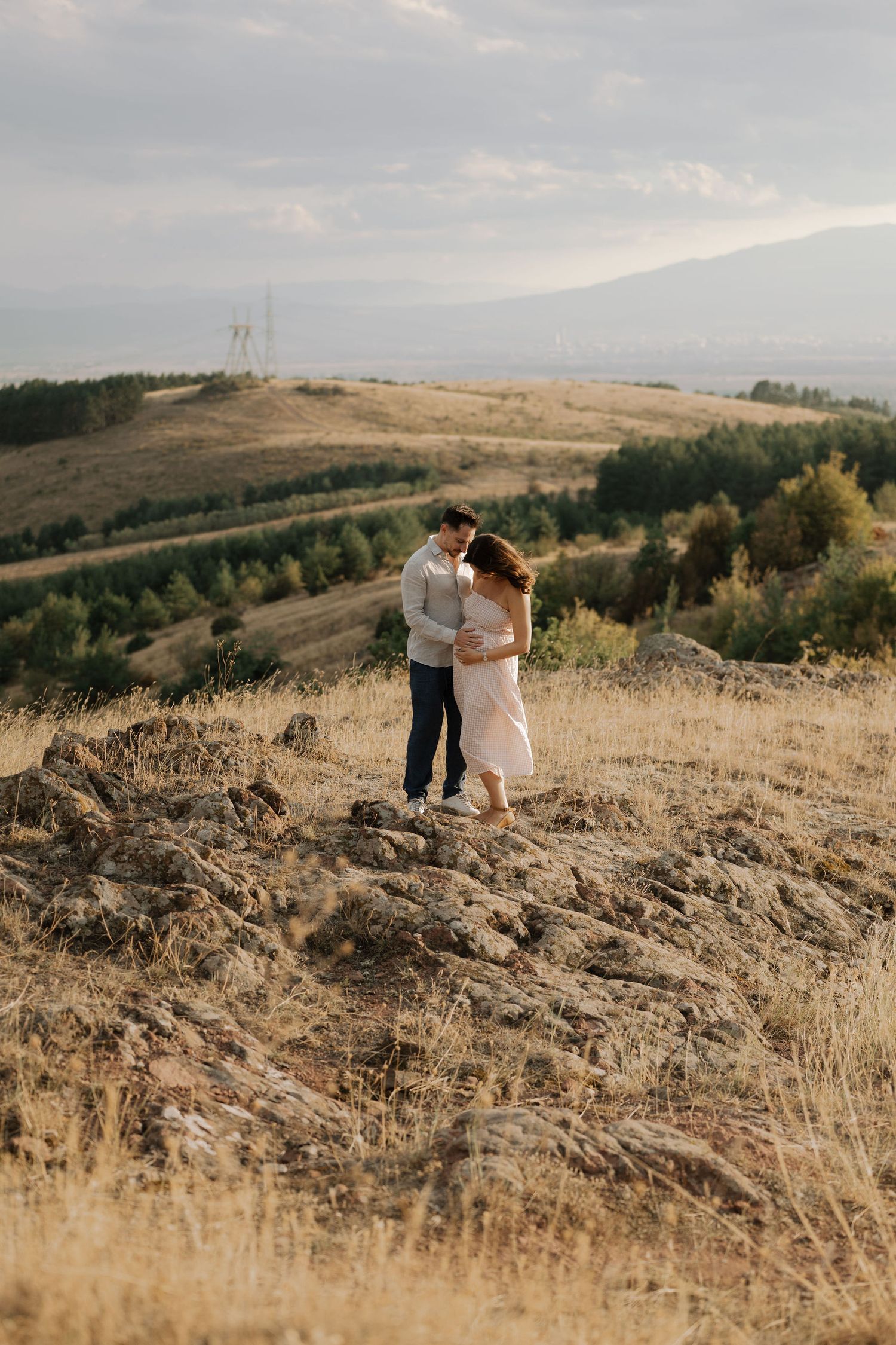 A couple embraces on a golden hillside at sunset while standing among dry grass and rolling terrain.