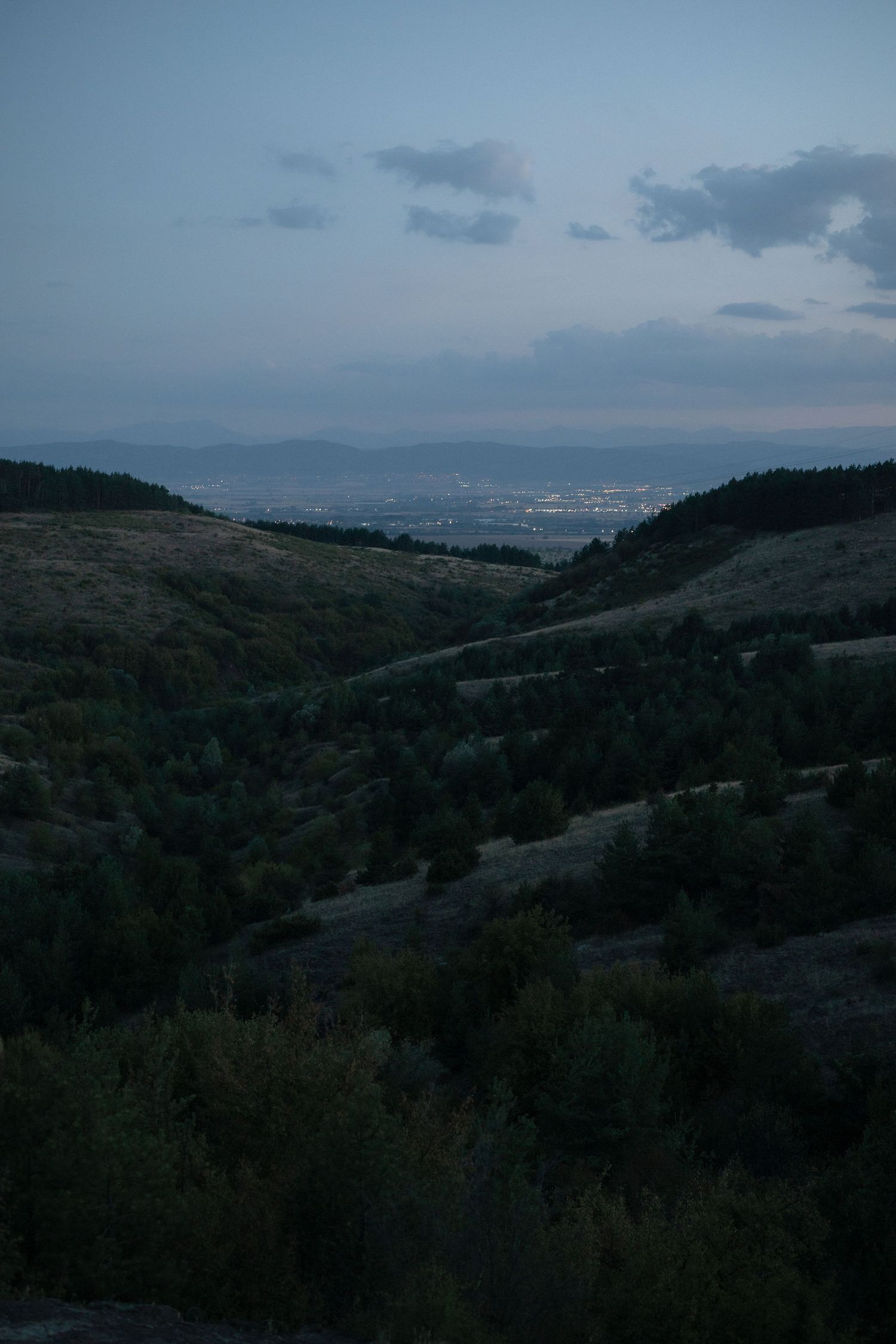 A moody twilight landscape overlooking a valley with rolling hills and distant mountains under a cloudy blue sky.