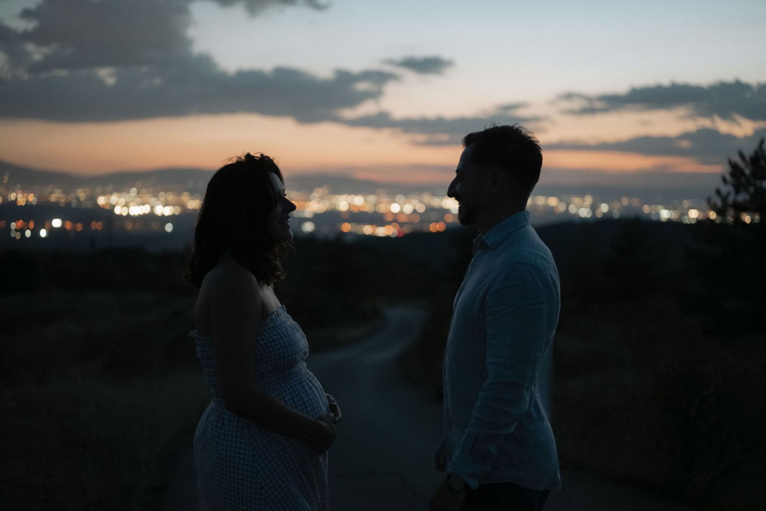 Two silhouettes stand together against a dusky cityscape with twinkling lights in the background at sunset.