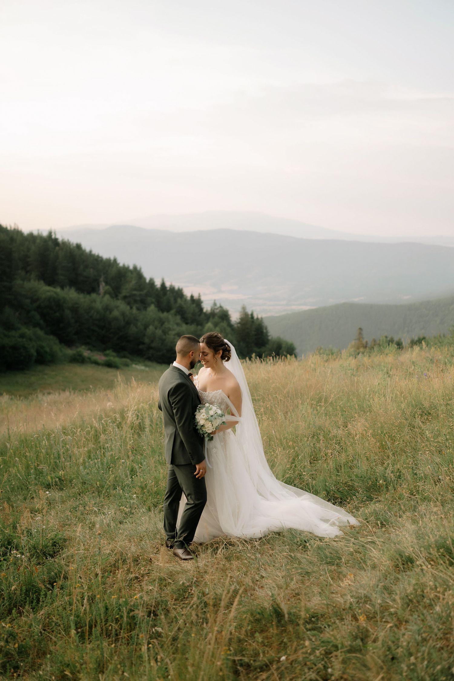 A romantic wedding couple embraces in a scenic mountain meadow during golden hour with rolling hills in the background.