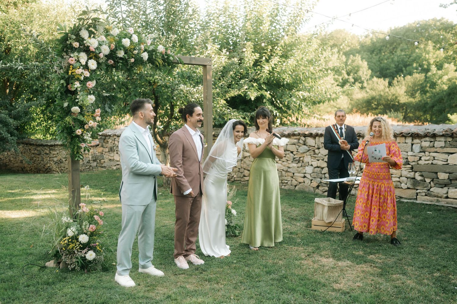 Wedding party stands in line during outdoor garden ceremony with floral arch and stone wall backdrop.