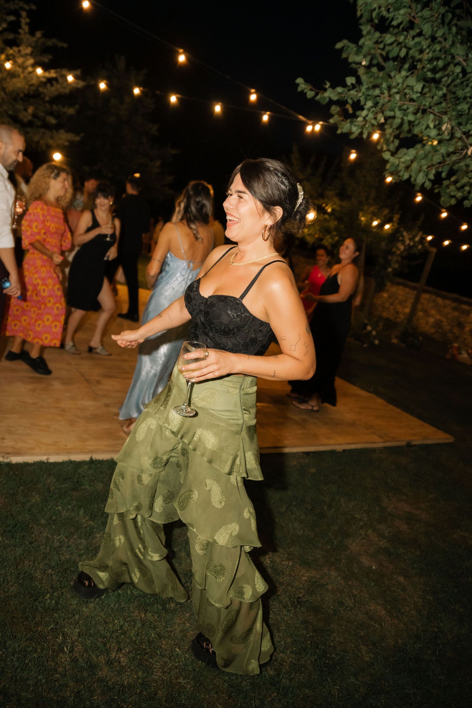 People dance together under string lights at an outdoor evening party on a grassy area.