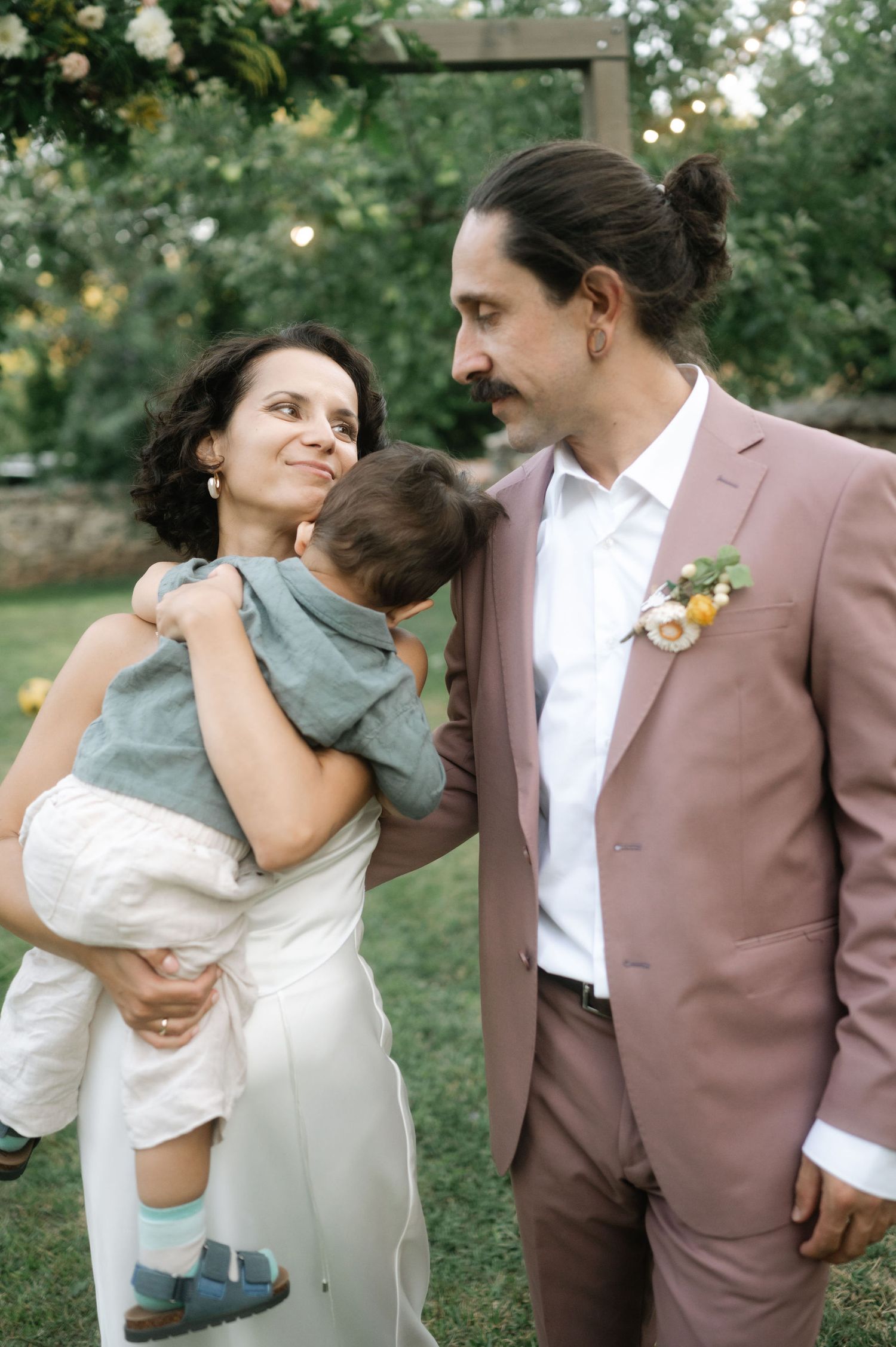 A couple shares tender moments with their child in an outdoor garden setting while wearing formal attire.