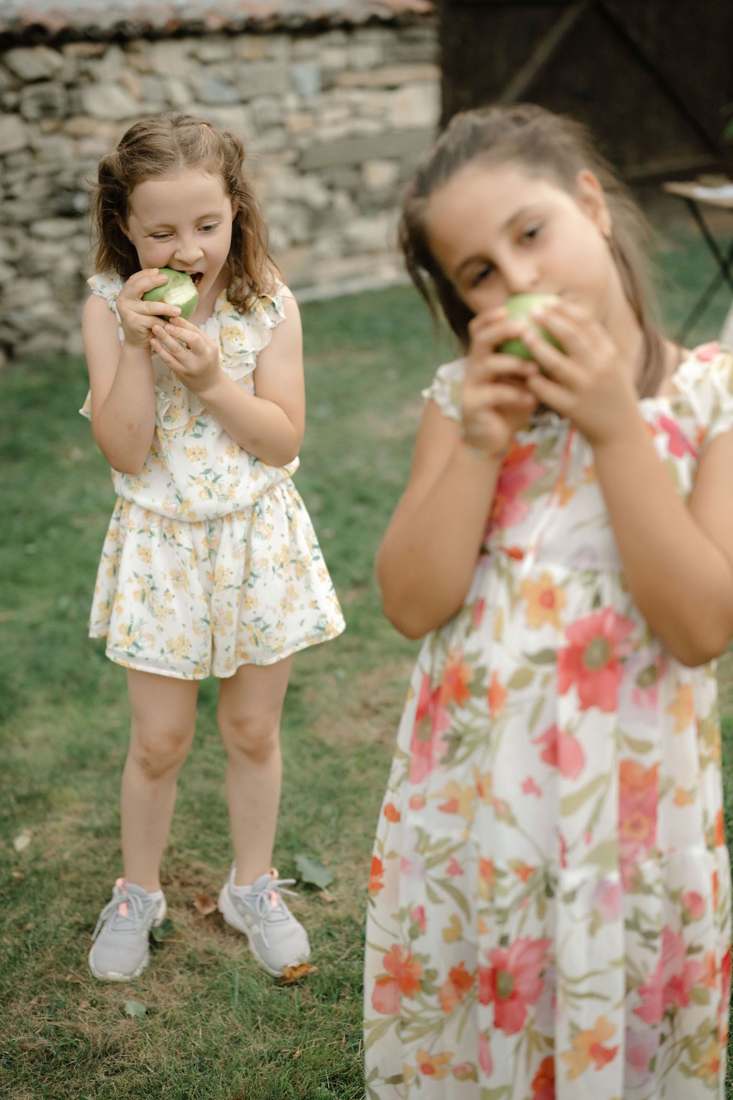 Two children in floral summer dresses enjoy snacks while playing outdoors on a sunny day.