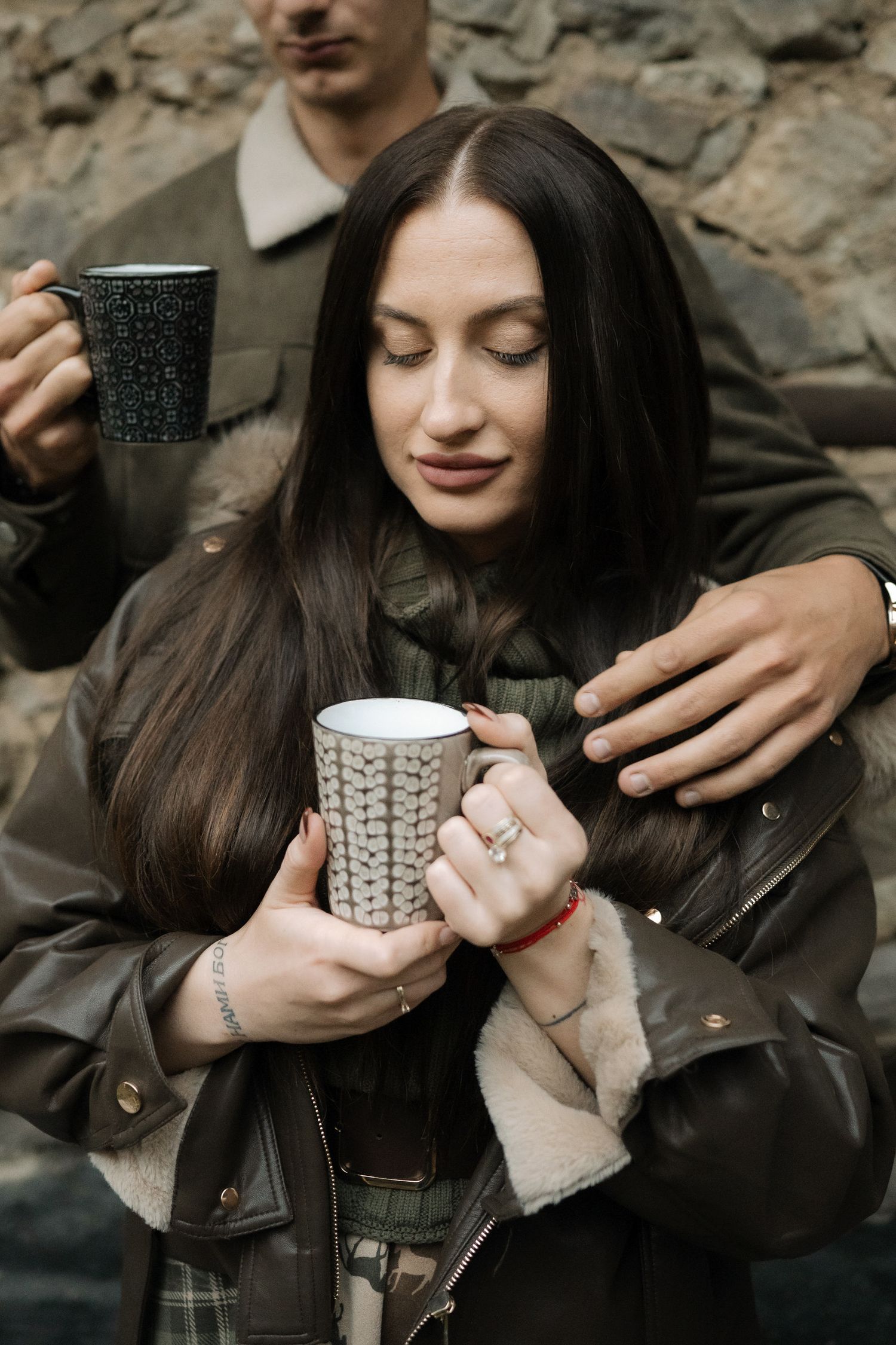 A couple in dark clothing shares intimate moments while holding knitted sweater coffee mugs outdoors.