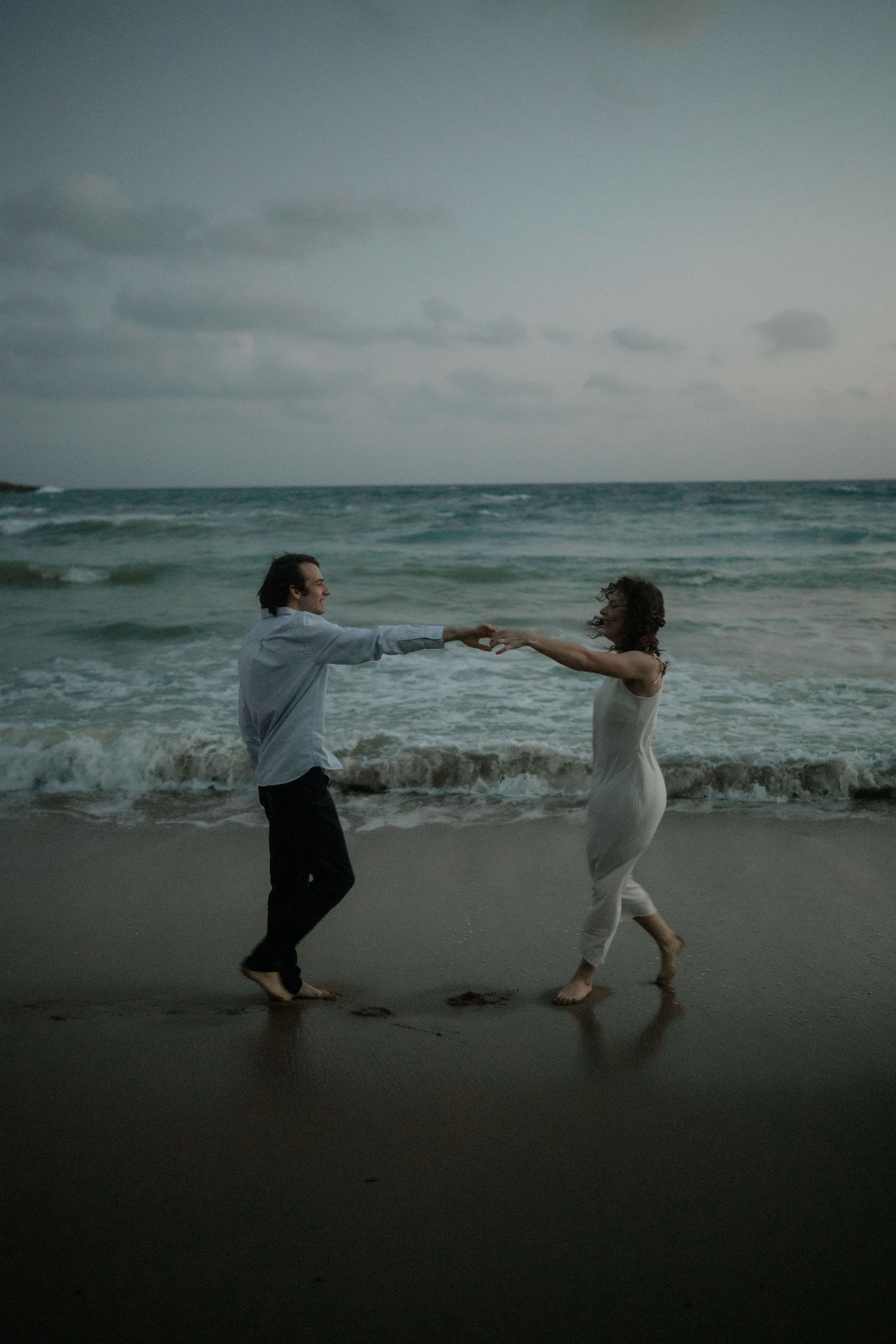 Couple dancing barefoot on a sandy beach at dusk, holding hands with ocean waves crashing behind them.