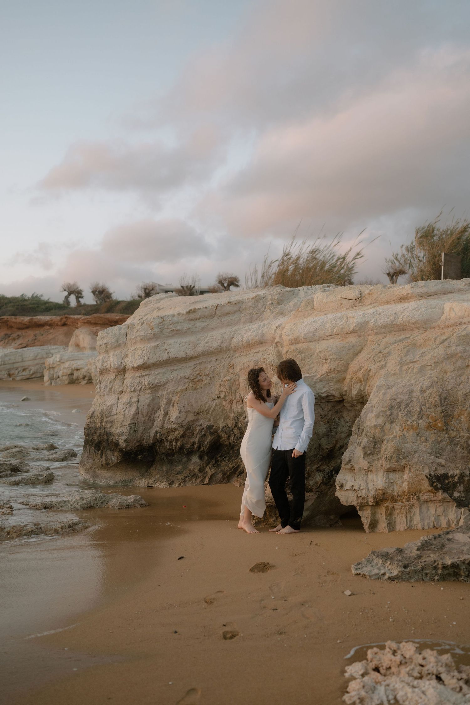 Romantic couple nearly kissing against rugged rock cliffs on a sandy beach at sunset.