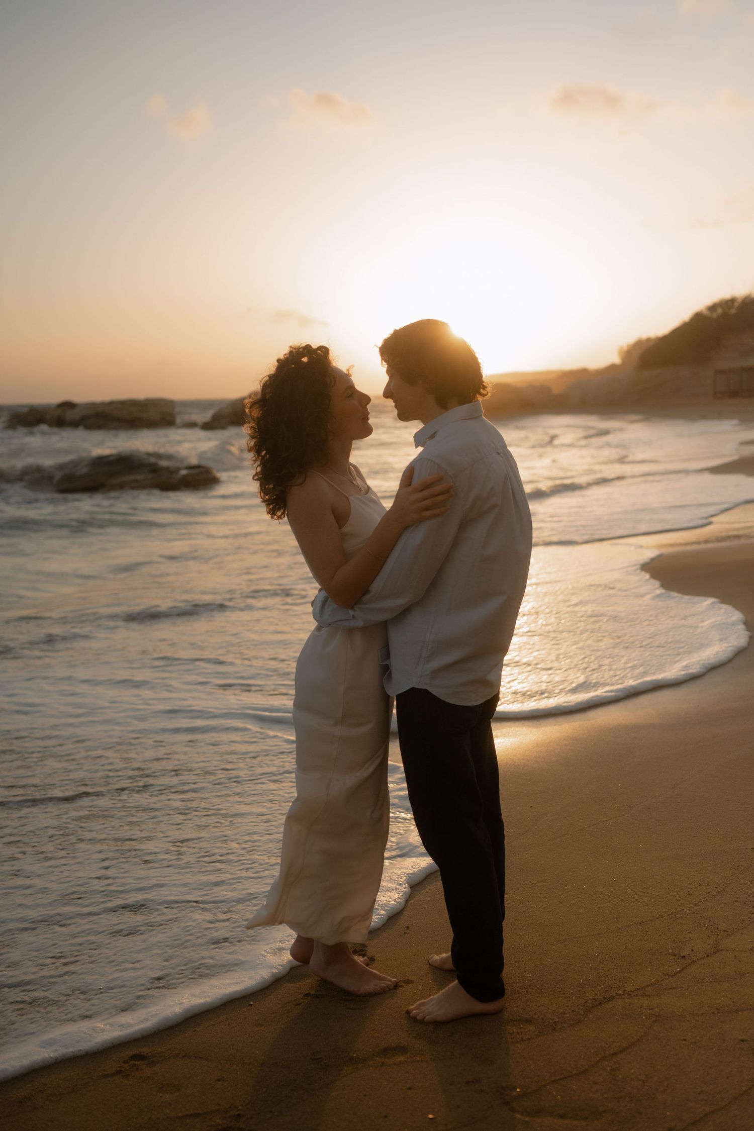 Romantic couple embracing on a sandy beach at golden sunset, waves gently washing ashore behind them.