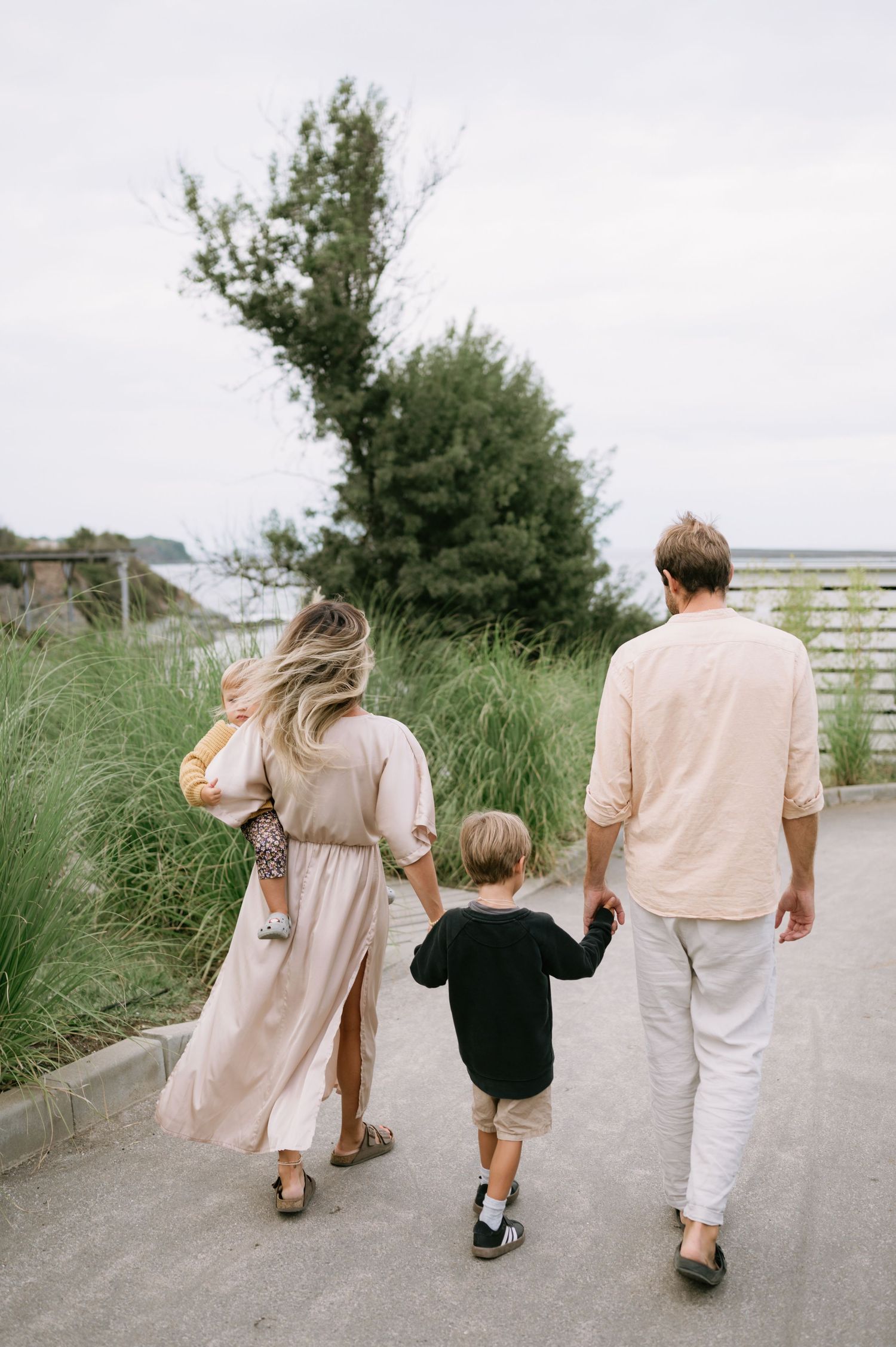 Family of four walking hand-in-hand on a coastal path surrounded by lush greenery on an overcast day.