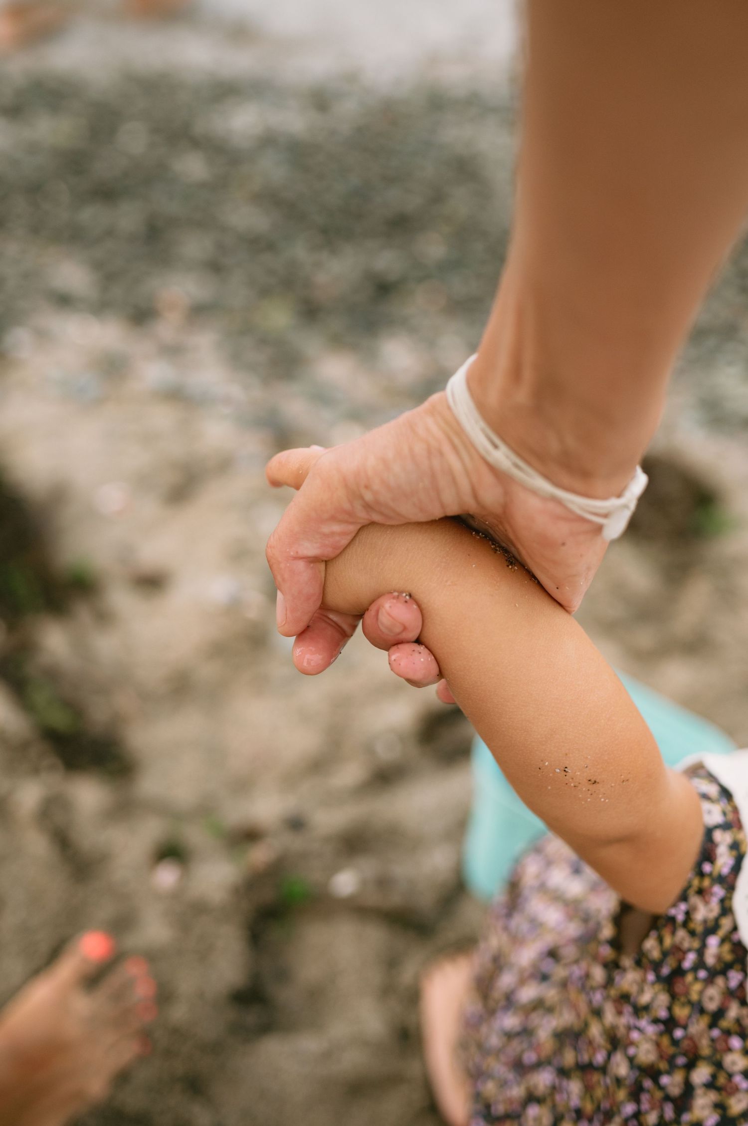 An adult hand with a white bracelet gently holding a young child's hand on a sandy beach.
