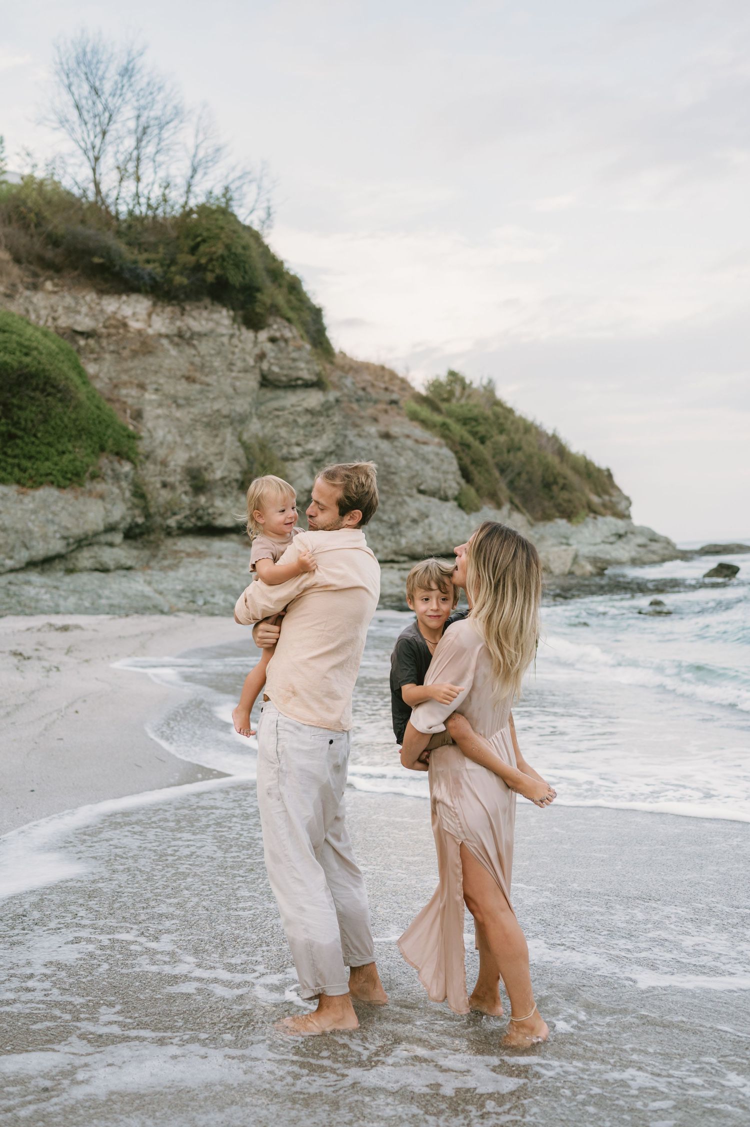 Family of four enjoying a beach day, parents holding young children near ocean waves with rocky cliffs in background.