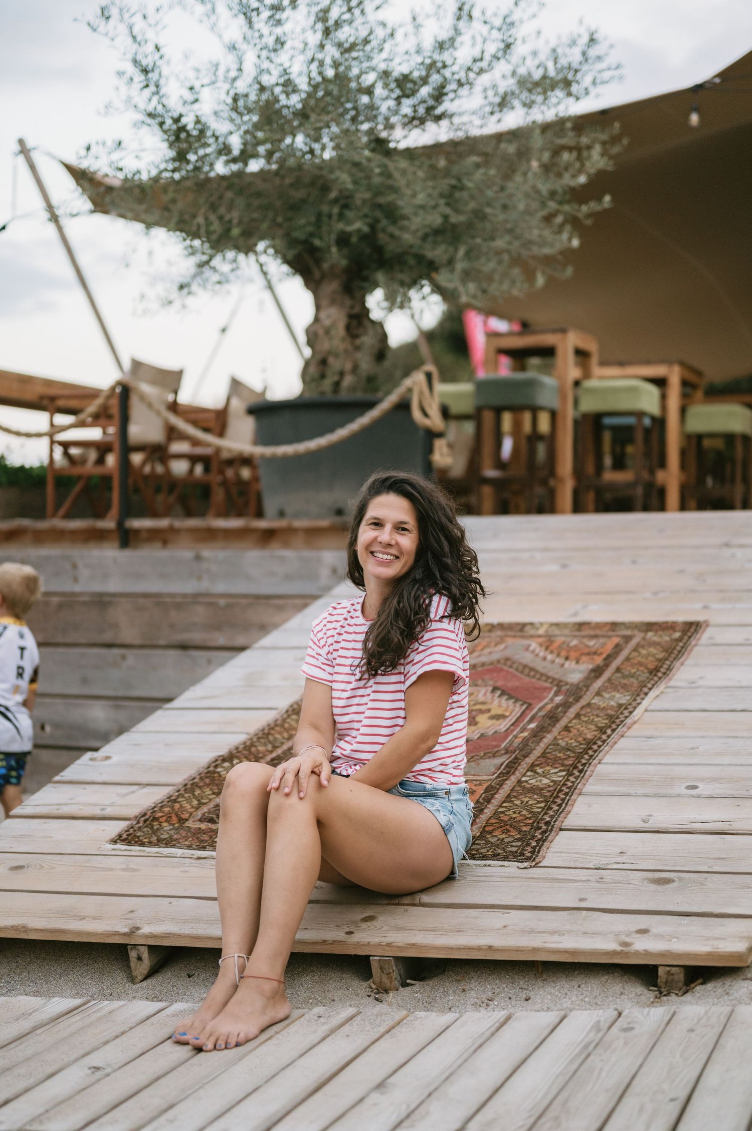 Smiling woman in striped tee and denim shorts sitting barefoot on wooden deck with patterned rug outdoors.