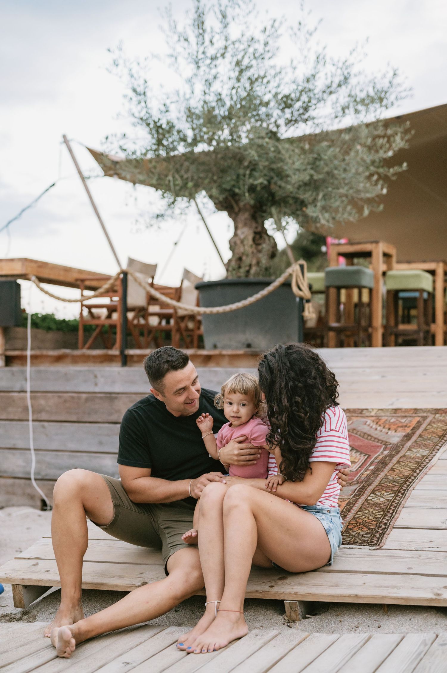 A young family with a toddler sitting on a wooden deck near an olive tree at an outdoor cafe.