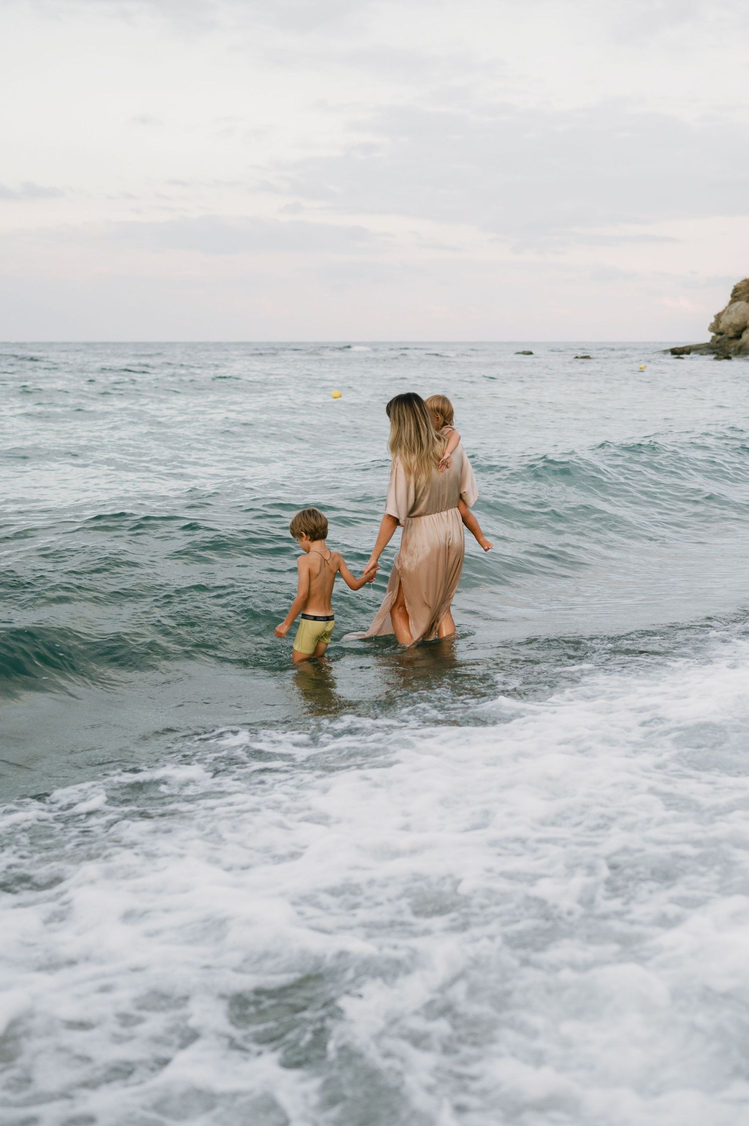 A mother and two children wade through ocean waves on a cloudy beach day.