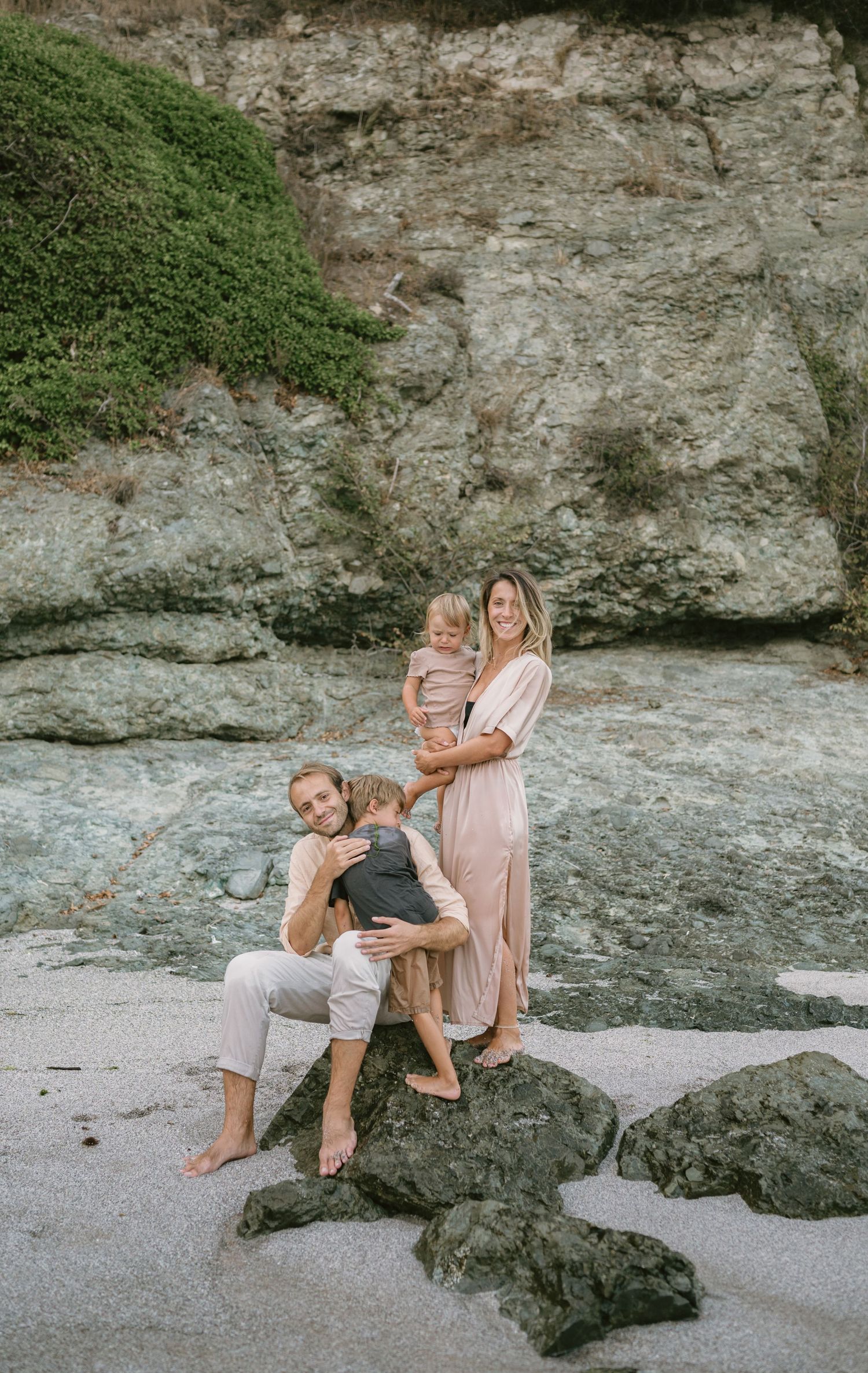 Family of four posing on rocky beach with cliff backdrop, mother in pink dress holding toddler, father seated with child.