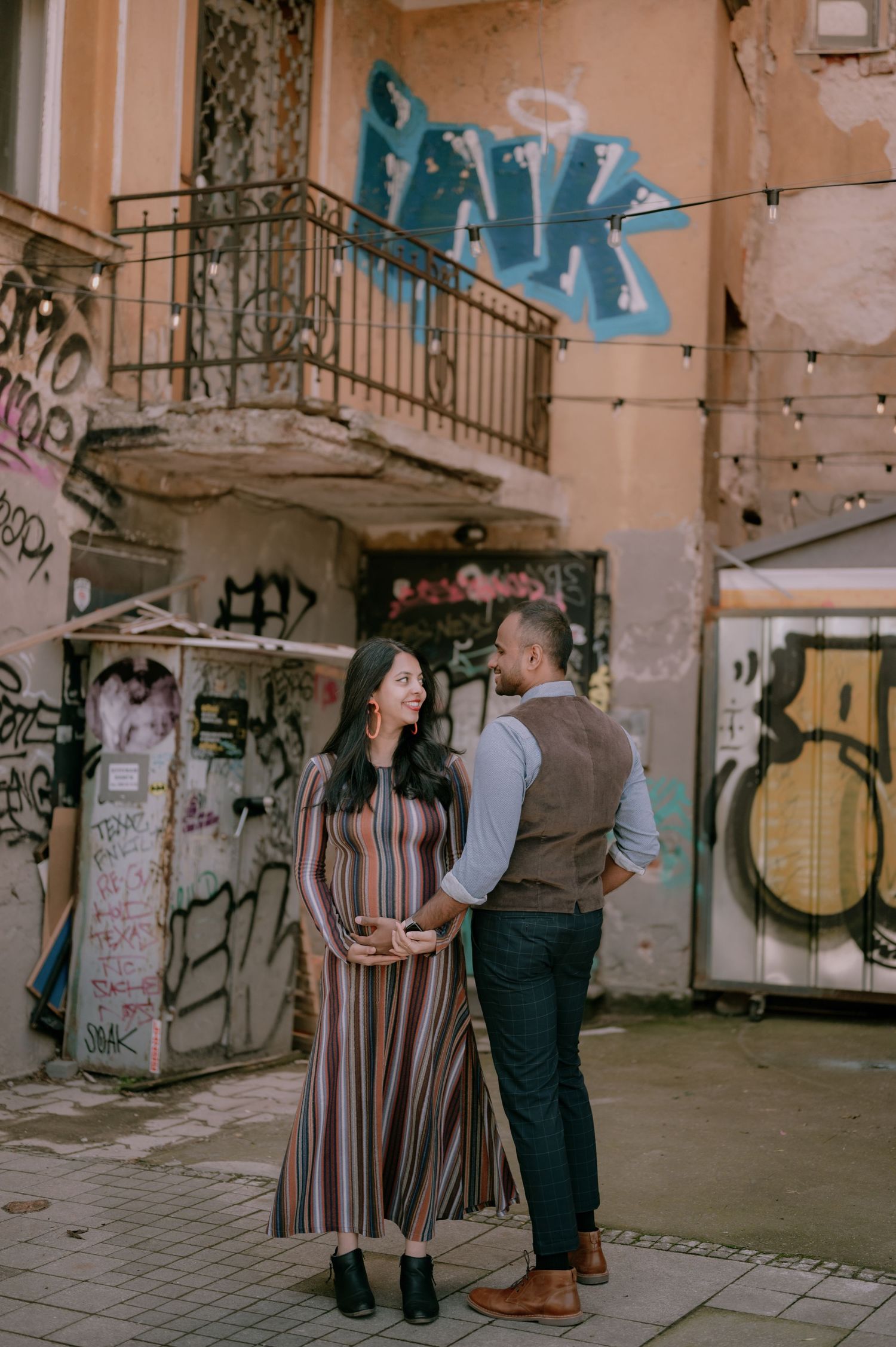 Pregnant woman in striped dress and man in vest share a tender moment in an urban alleyway with colorful graffiti.