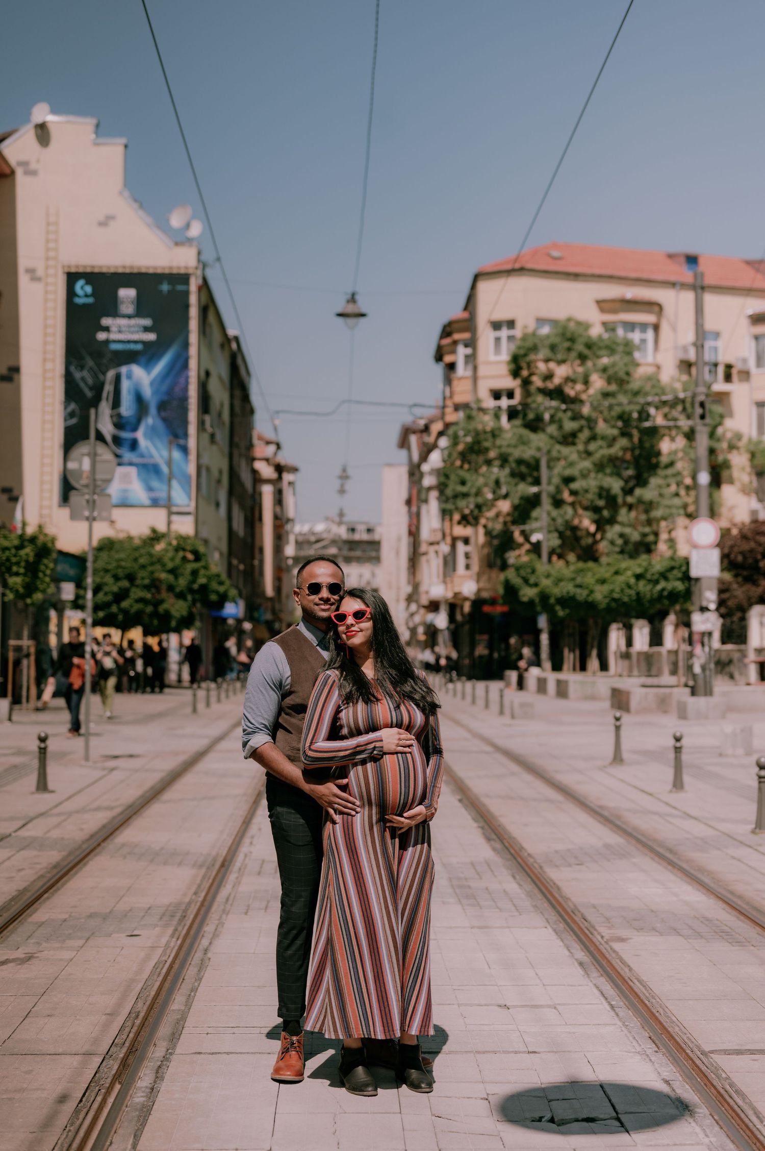 Pregnant couple posing on urban tram street, woman in striped maxi dress, man in gray shirt, city buildings behind.